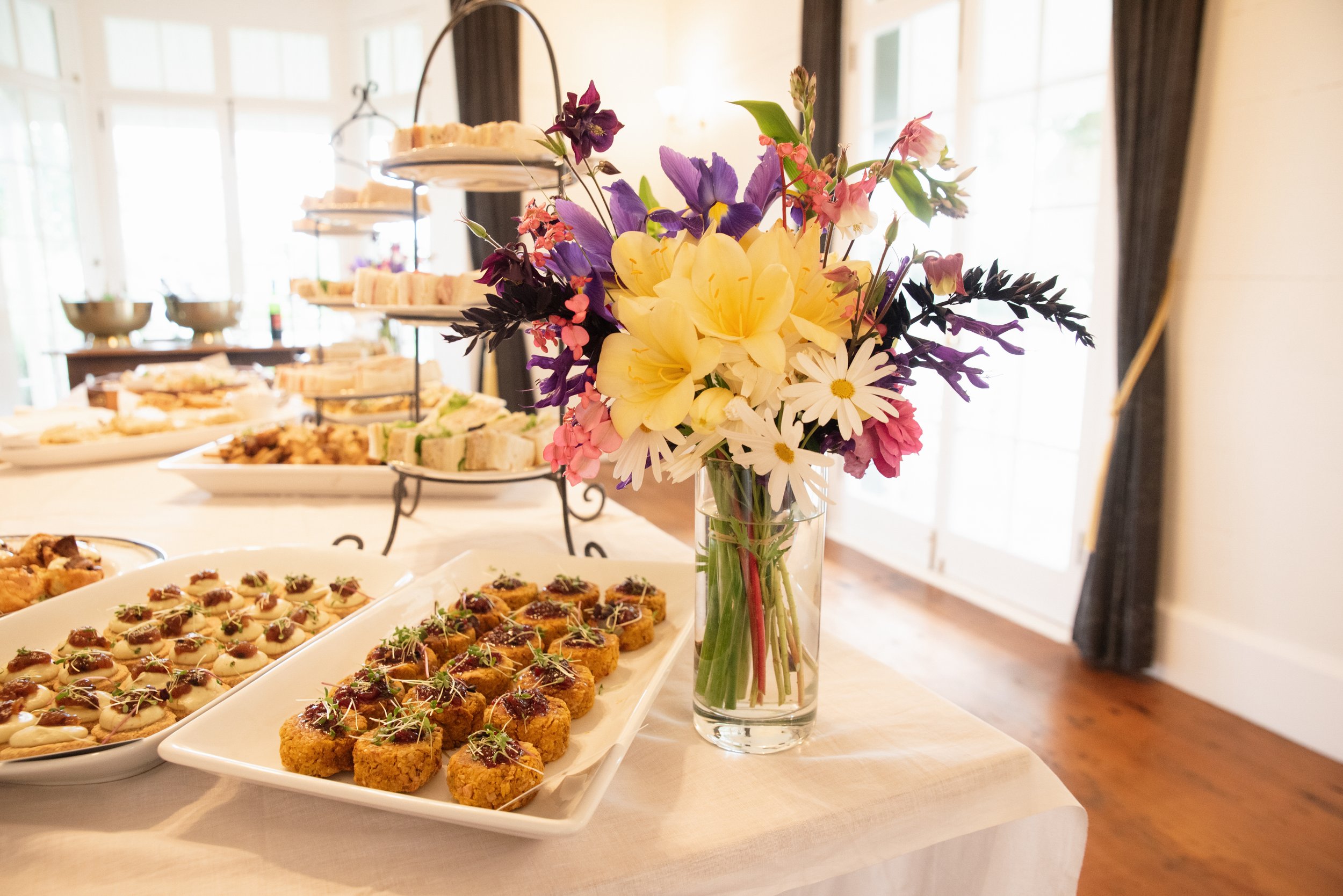 A bouquet of colorful flowers in a glass vase on a table, with a spread of assorted appetizers and finger foods in the background, set in a bright room with large windows and dark curtains.