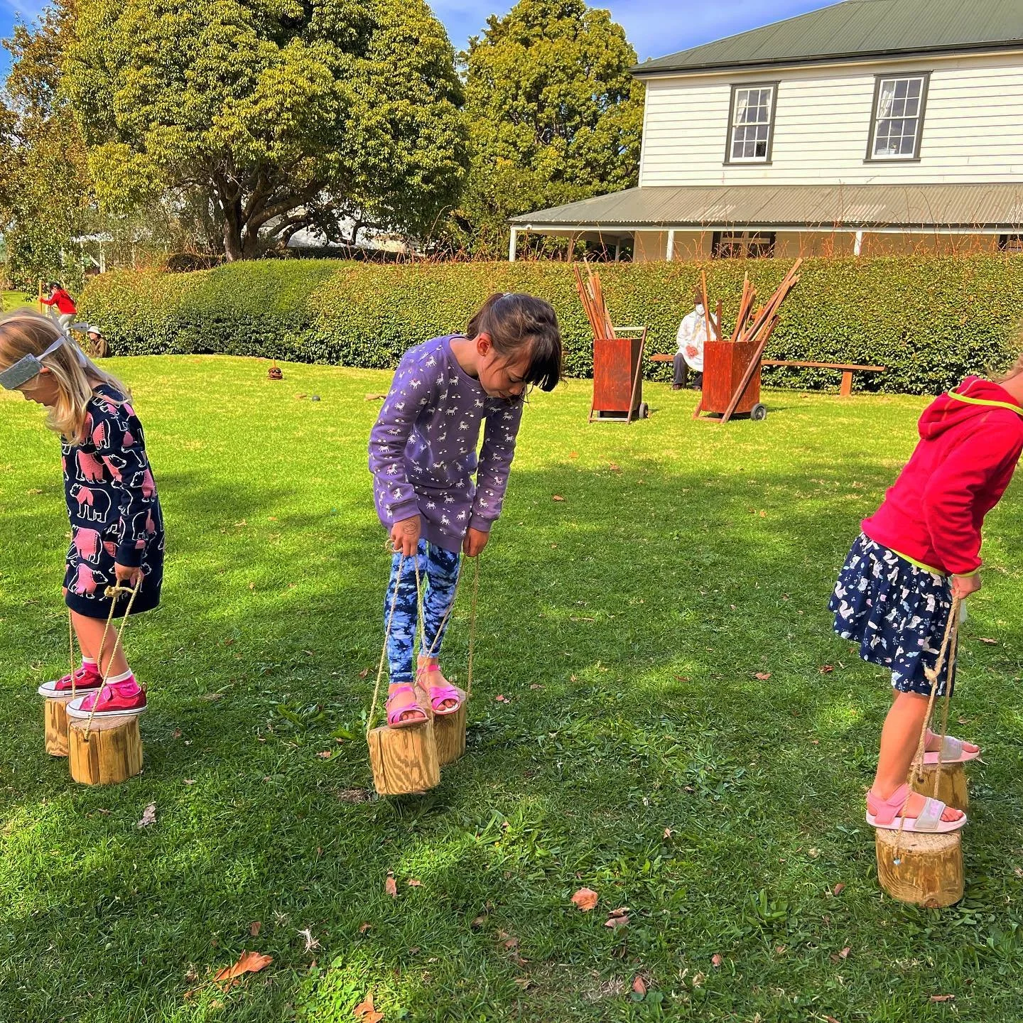 Three children walking on wooden stumps tied with ropes on a grassy field outdoors.