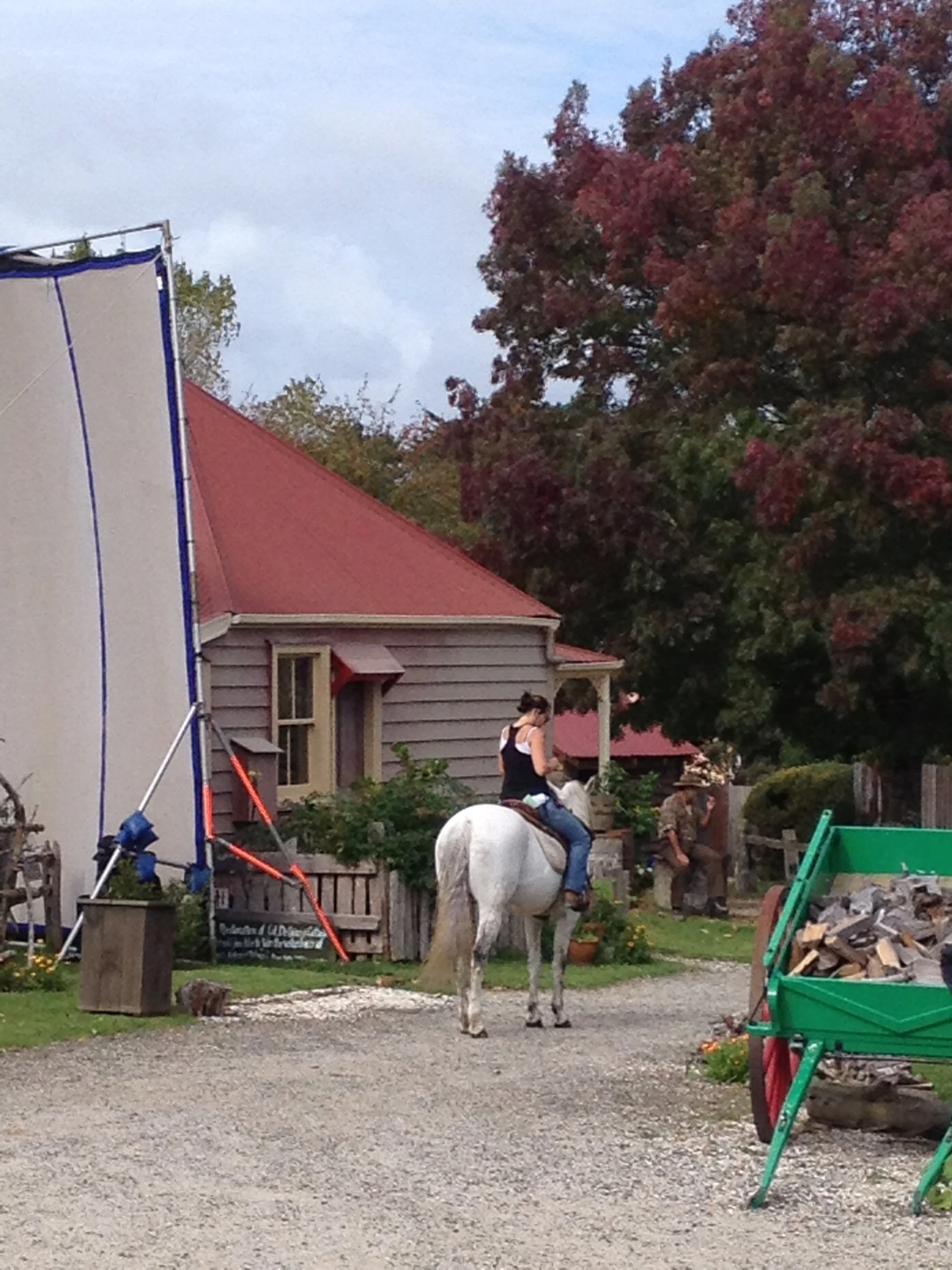 Woman riding a white horse in a rustic outdoor setting with a red-roofed house, trees with red leaves, a green wheelbarrow filled with firewood, and a man standing nearby.
