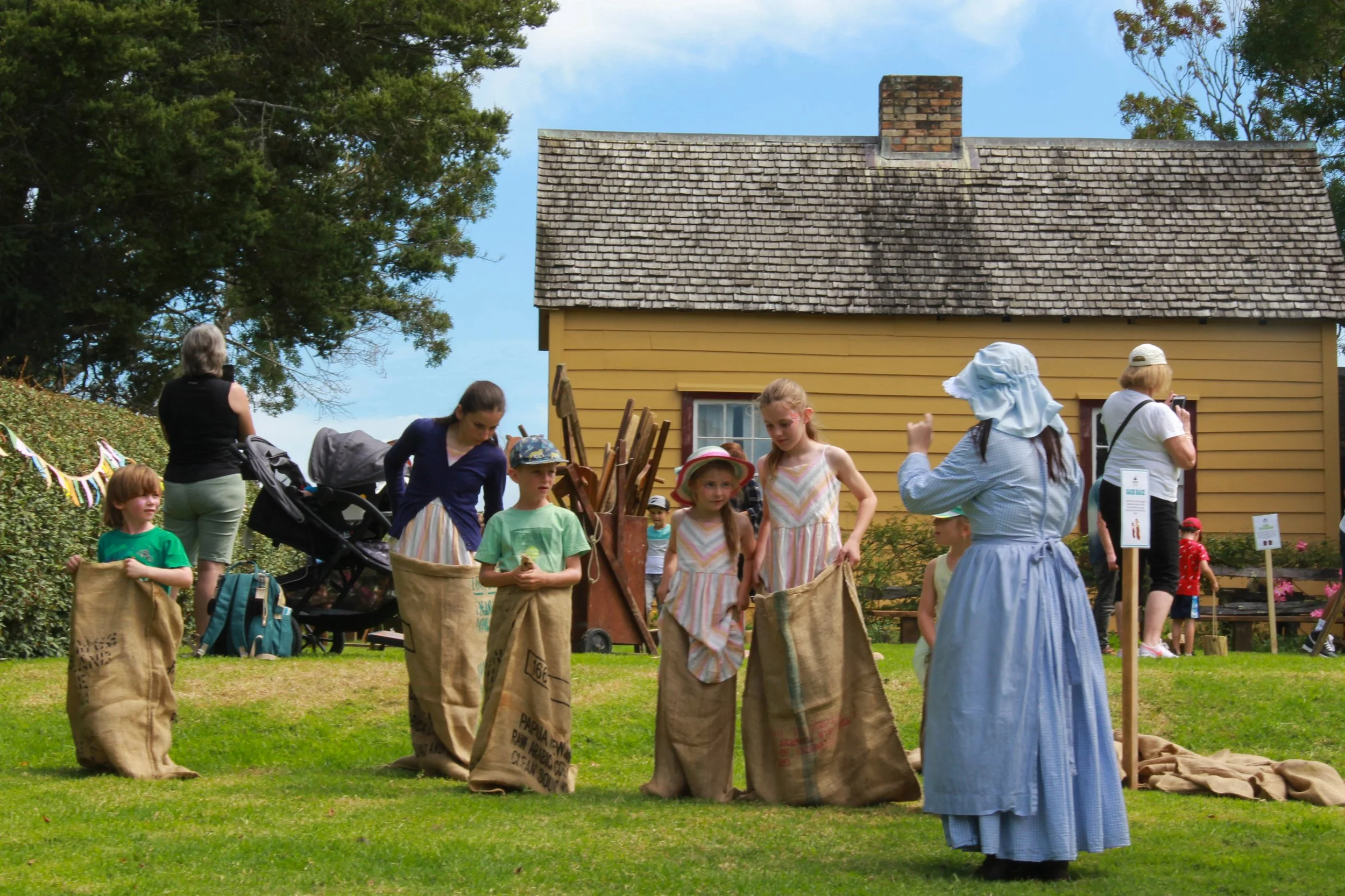 Children playing sack races outdoors on a sunny day with a yellow house in the background.