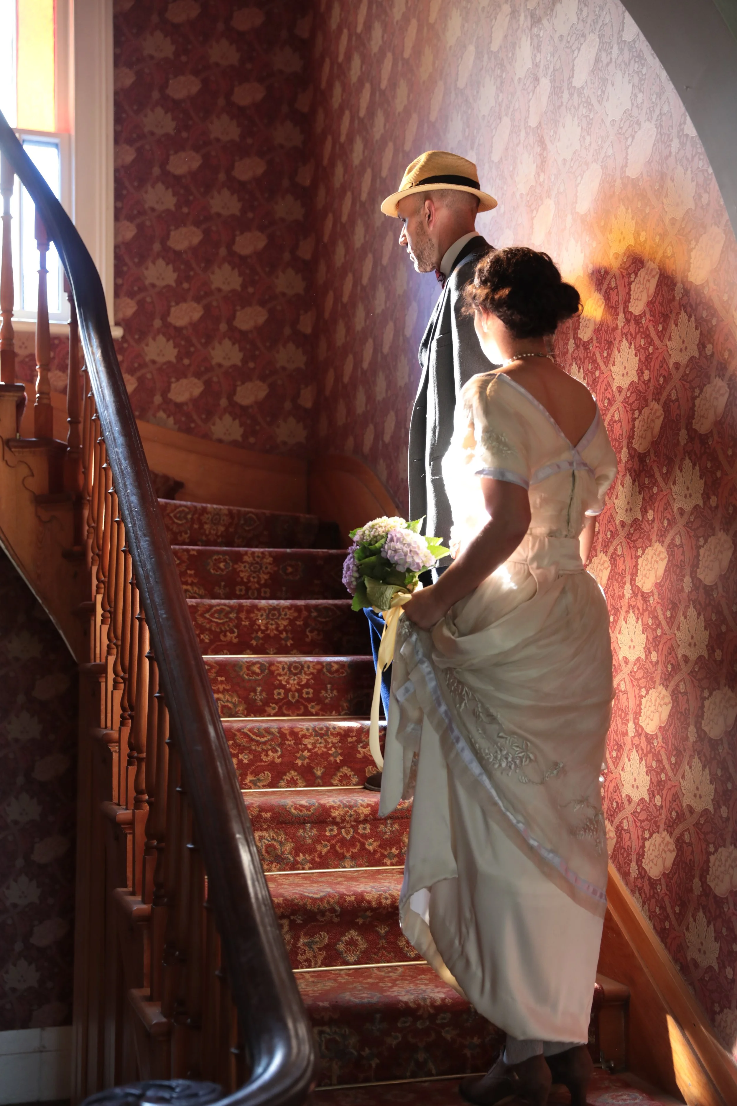 A couple dressed in wedding attire descending a red-carpeted staircase, with sunlight shining on them. The man is wearing a hat and a suit, while the woman is holding a bouquet of flowers.