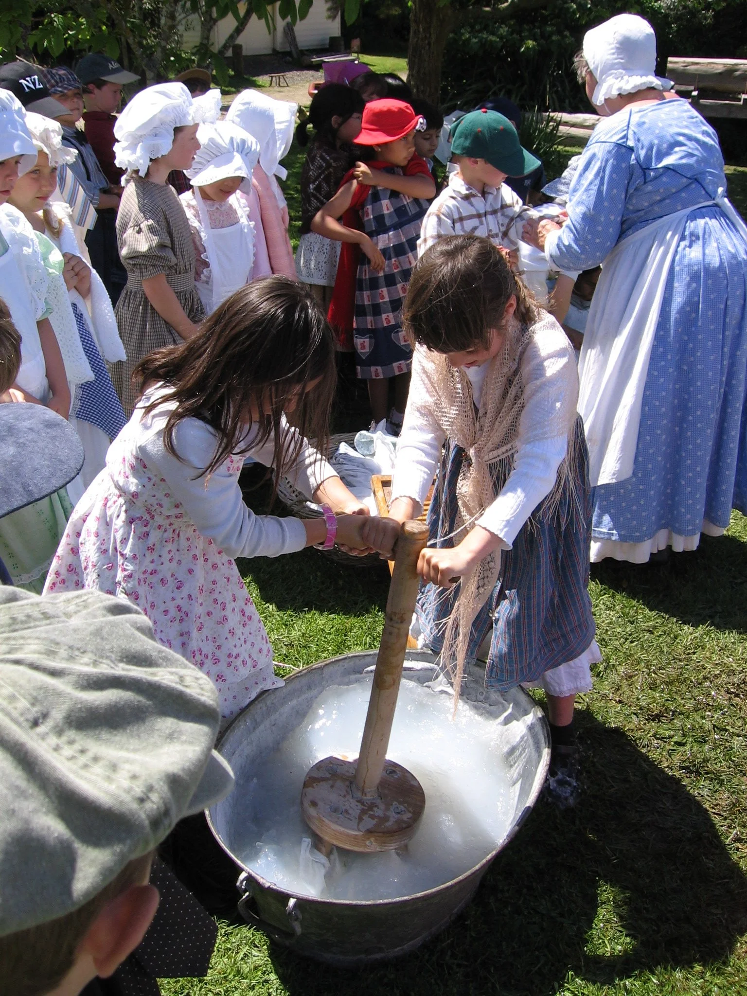 Children dressed in historical costumes participating in a cotton candy or similar treat activity outdoors under the supervision of a woman in period clothing.