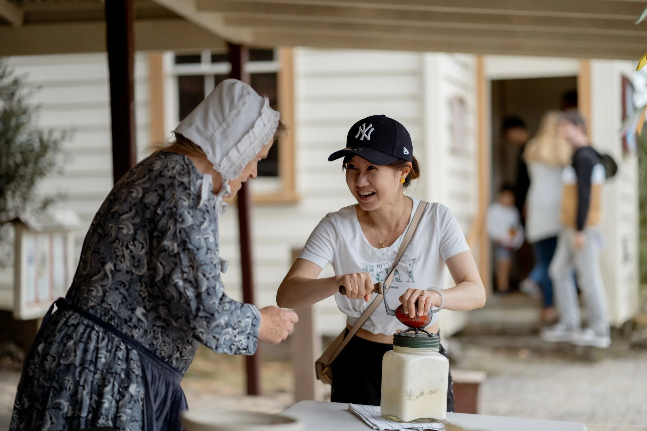Two women smiling while using a vintage butter churn outside a building with onlookers in the background.