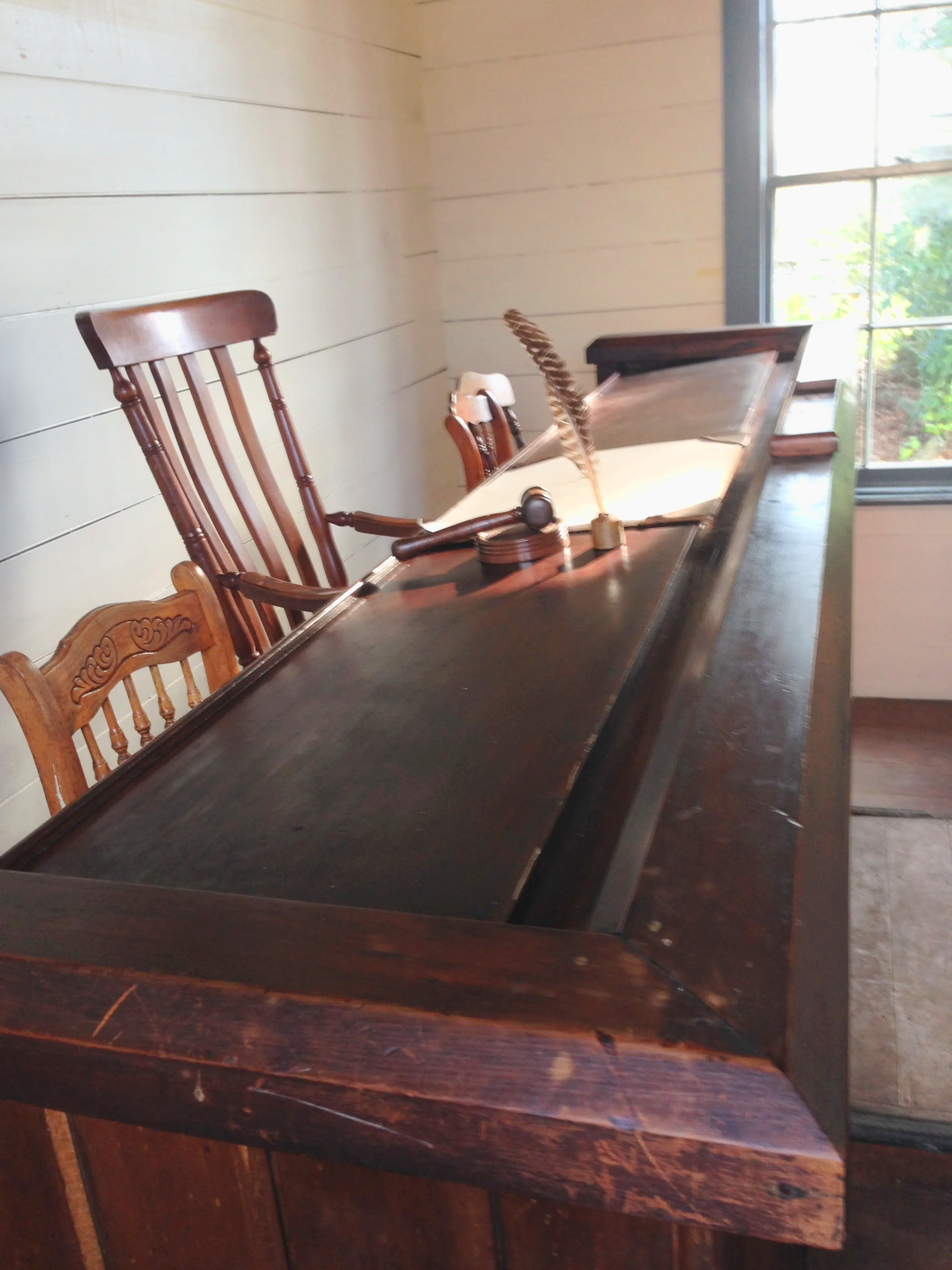 View of a wooden writing desk with a feather quill and inkwell, and several wooden chairs nearby, next to a large window with greenery outside.
