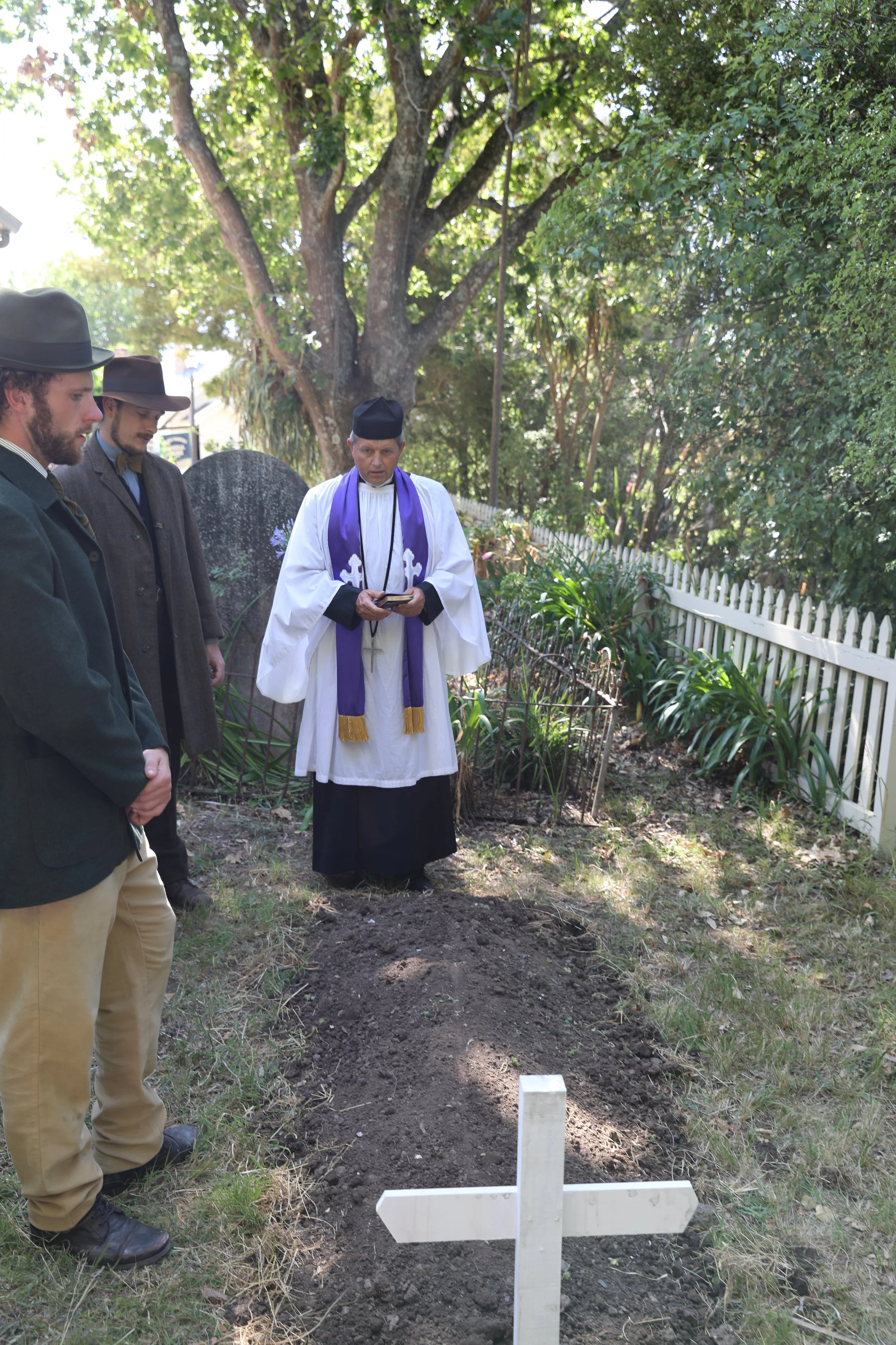 Three men and a priest standing near a grave marked by a white cross in a garden with trees and bushes.