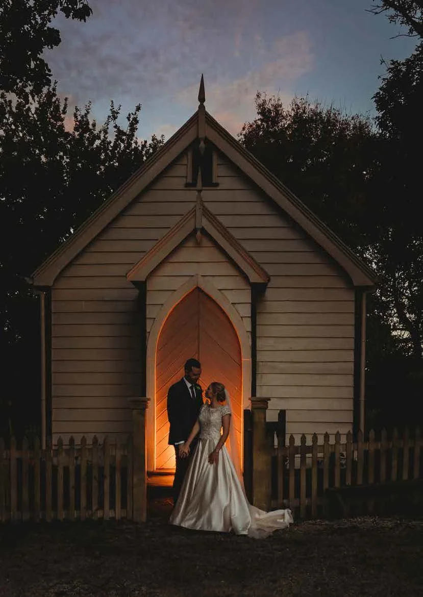 A bride and groom standing in front of a small, white wooden chapel with a pointed roof and illuminated doorway at dusk.