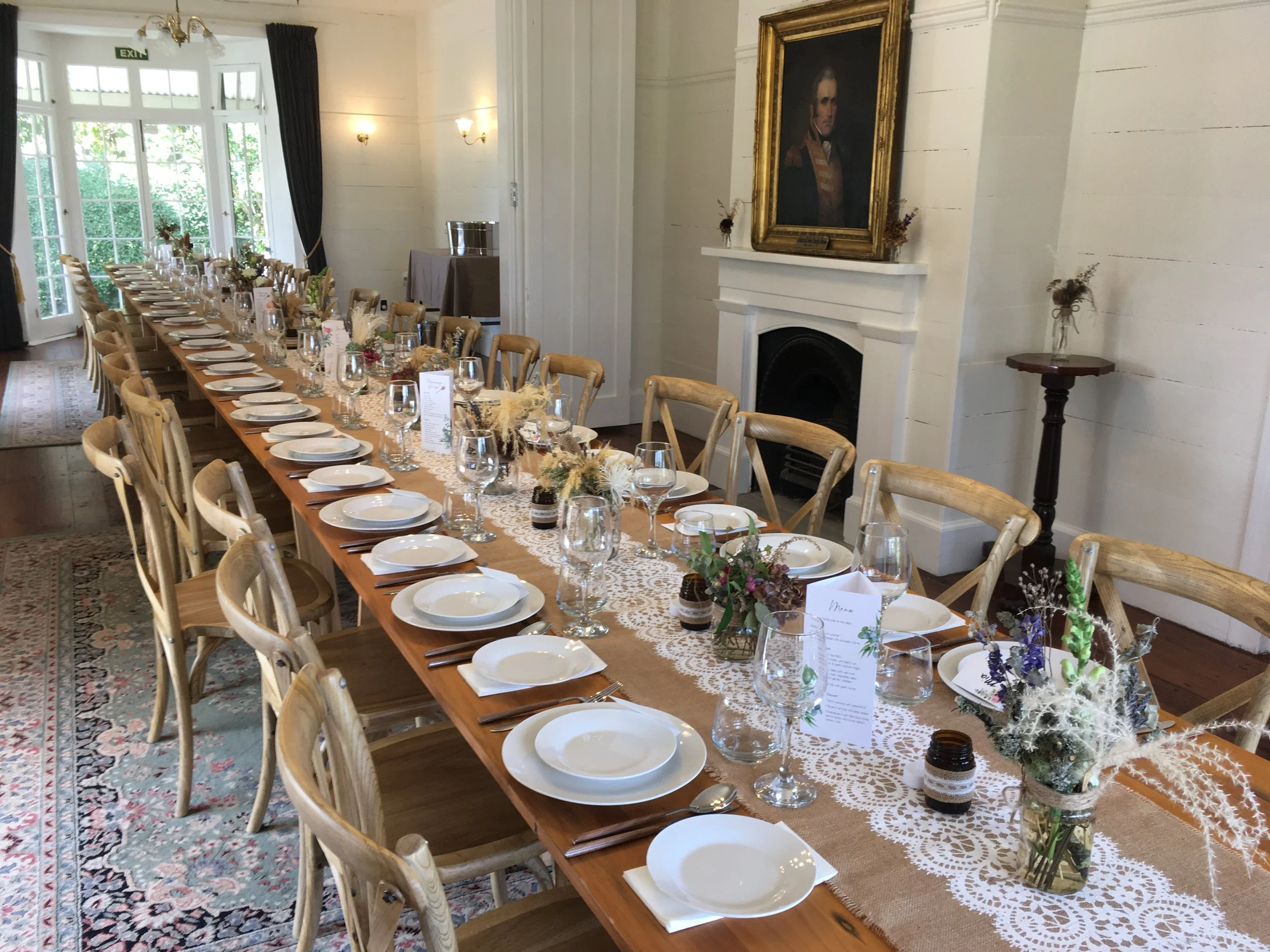 A long wooden dining table set for a formal event with white plates, wine glasses, and floral centerpieces, in a room with large windows, dark curtains, and a portrait painting of a man in historical attire hanging above a white fireplace.