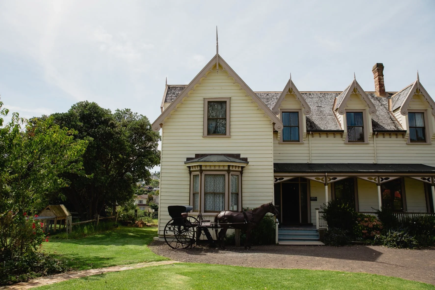 Vintage yellow wooden house with bay windows, steep gable roofs, and decorative trim; horse-drawn carriage parked outside; garden with trees and lawn.