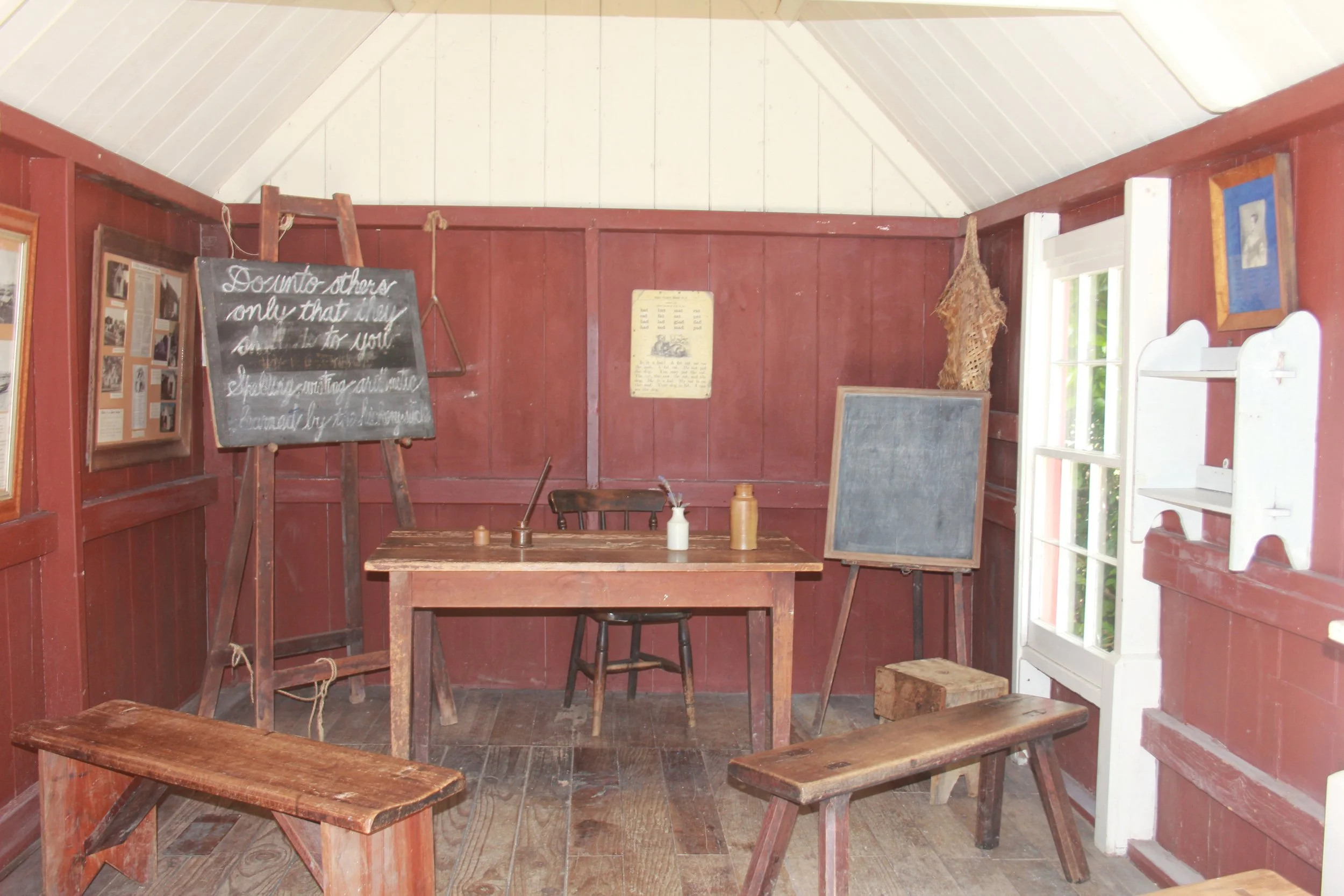 Interior of a rustic log cabin room with wooden tables, benches, a blackboard, and framed photographs on the red wooden walls, with a window on the right side.