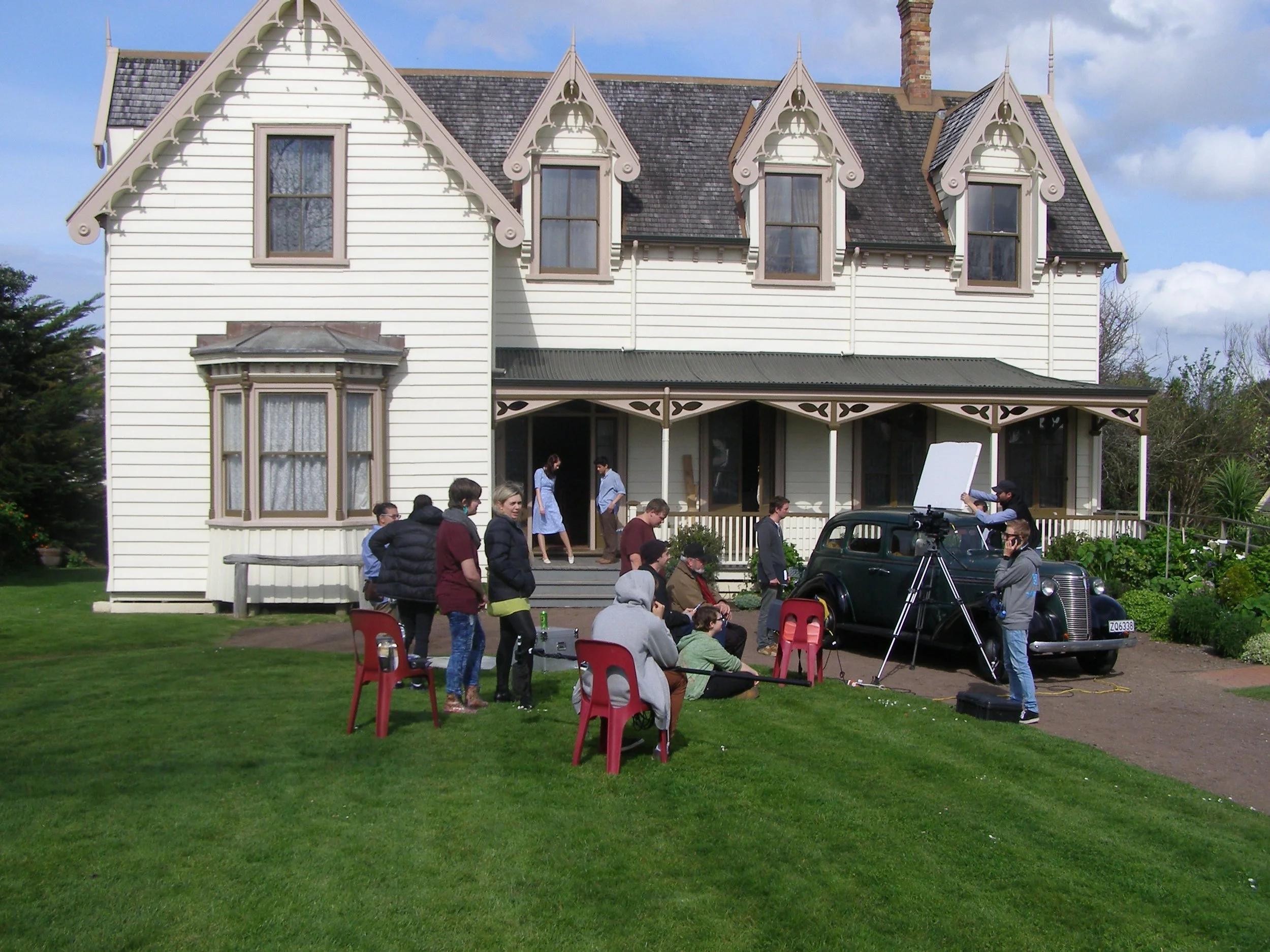 Film crew filming outside an ornate Victorian-style house with several crew members, actors, and vintage car on a green lawn.