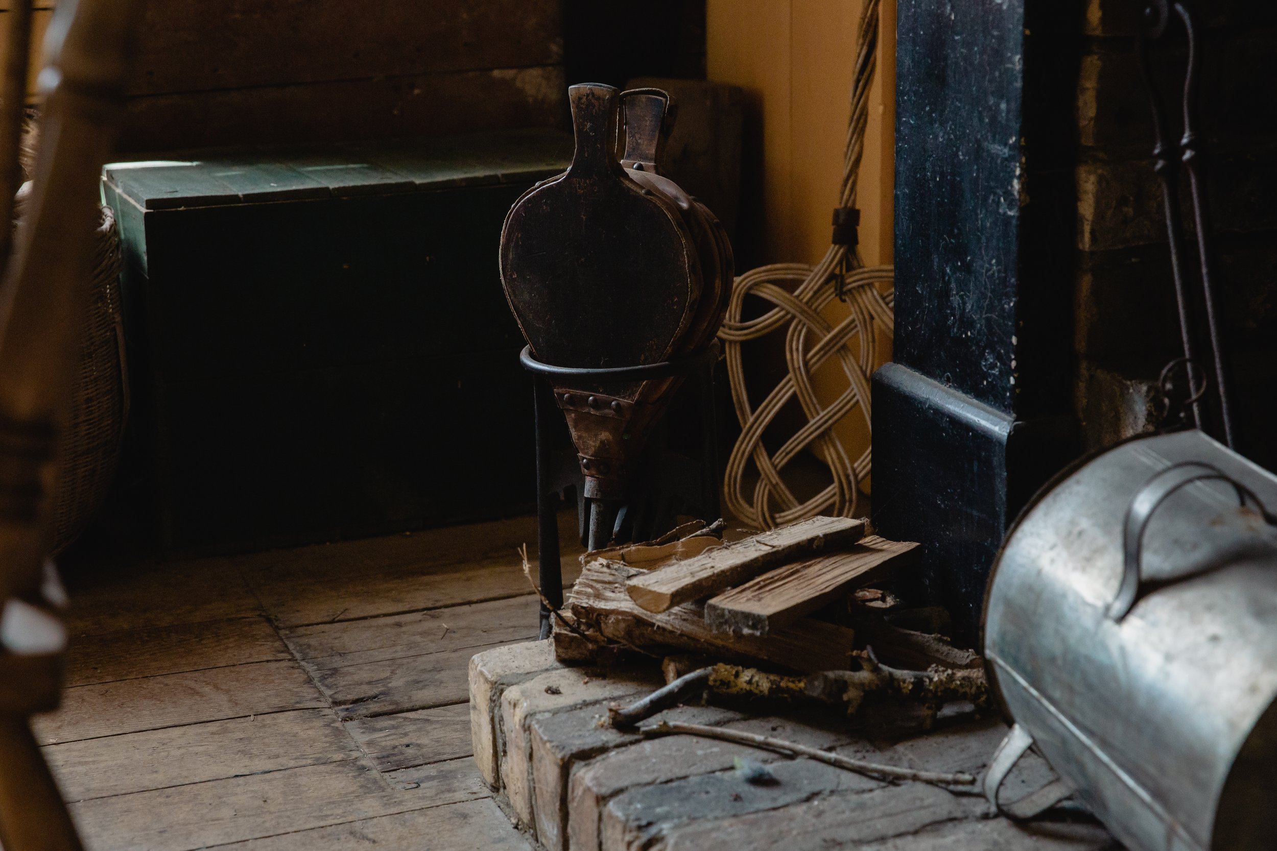A rustic interior scene with a pile of firewood, an old metal container, and a small black chest on a wooden floor, with a vintage bottle-shaped object and woven wicker in the background.
