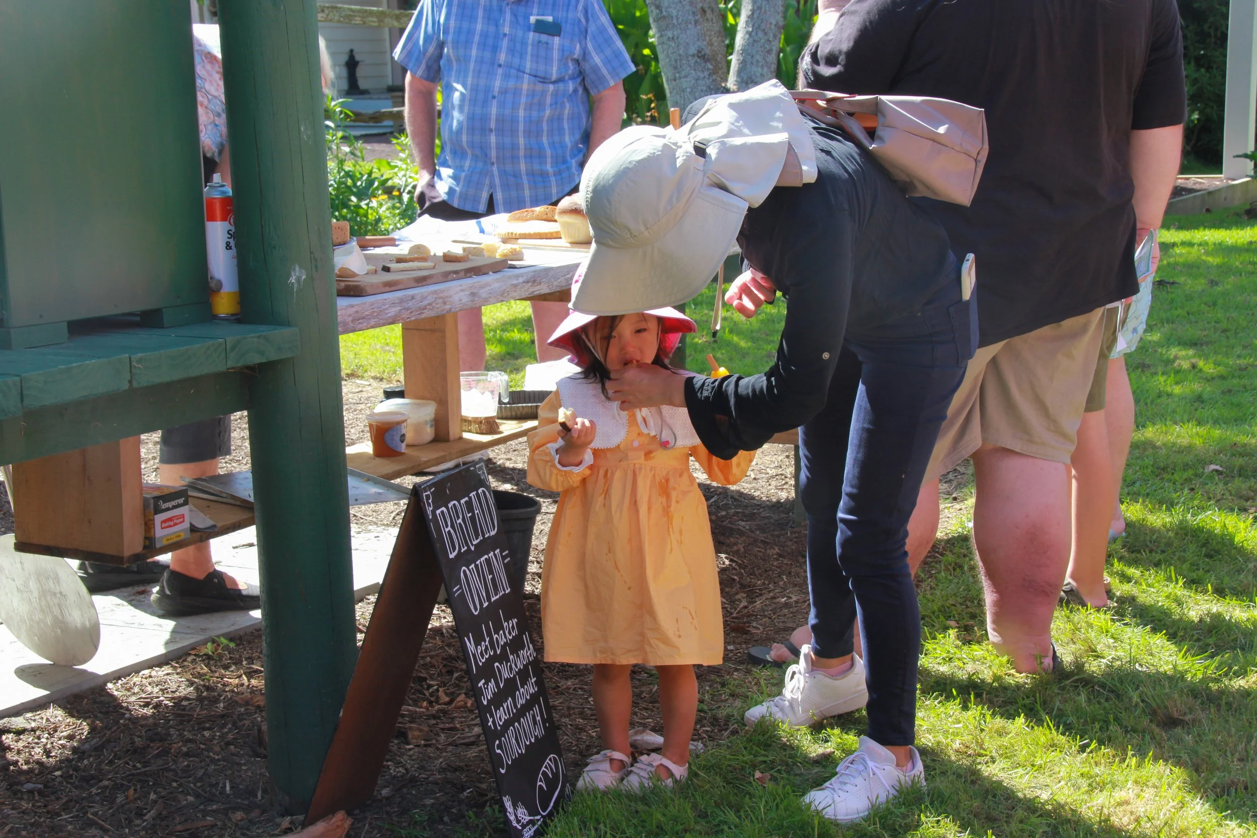 A young girl in a yellow dress and wide-brimmed hat is being fed a snack by an adult woman at an outdoor bake sale or market. There are other people in the background near a table with baked goods, and a blackboard sign nearby with chalk writing.