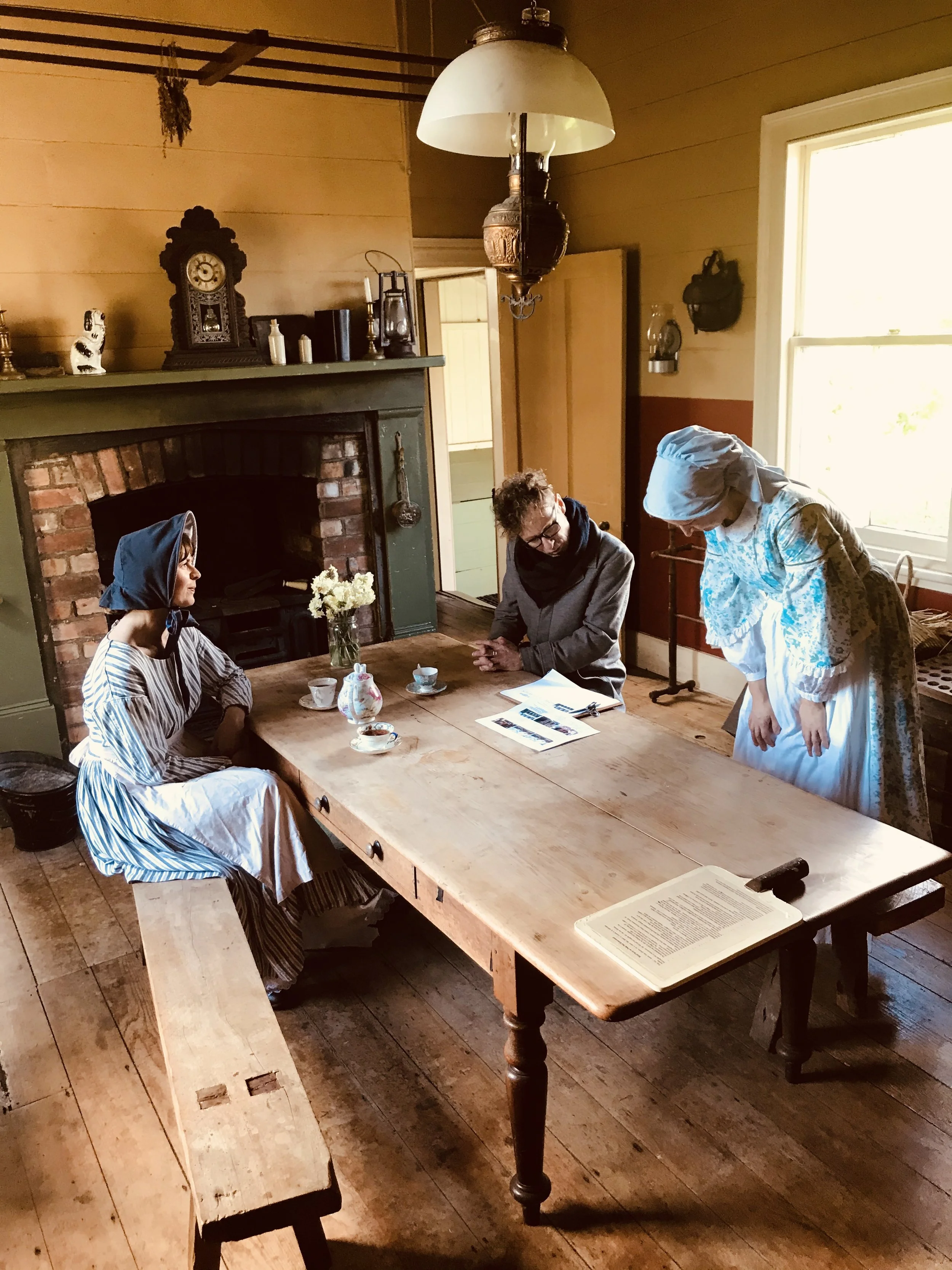Three people dressed in historical clothing sit at a wooden dining table in a cozy, rustic room with a brick fireplace, antique clock, and a large window letting in natural light.