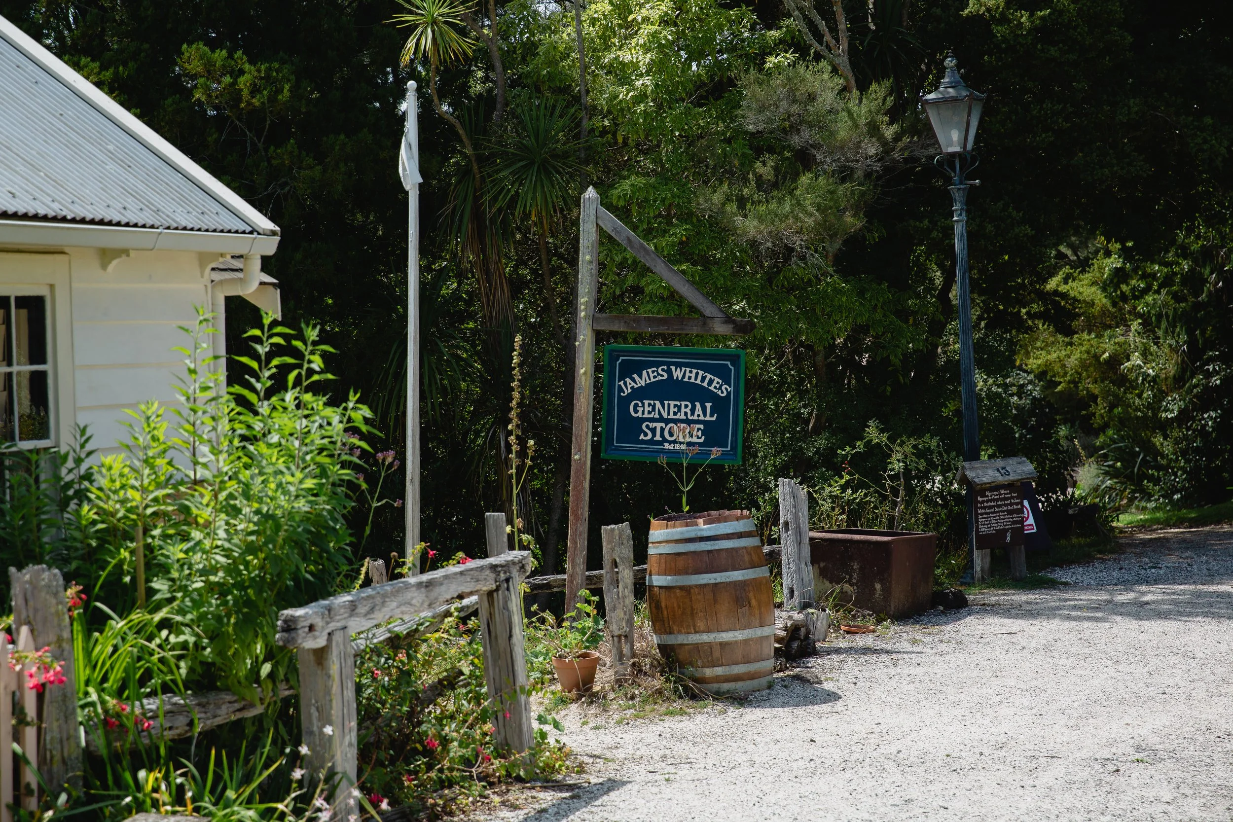 Sign for James Whites General Store outside a building surrounded by plants, an old wooden fence, a barrel, and a vintage street lamp.