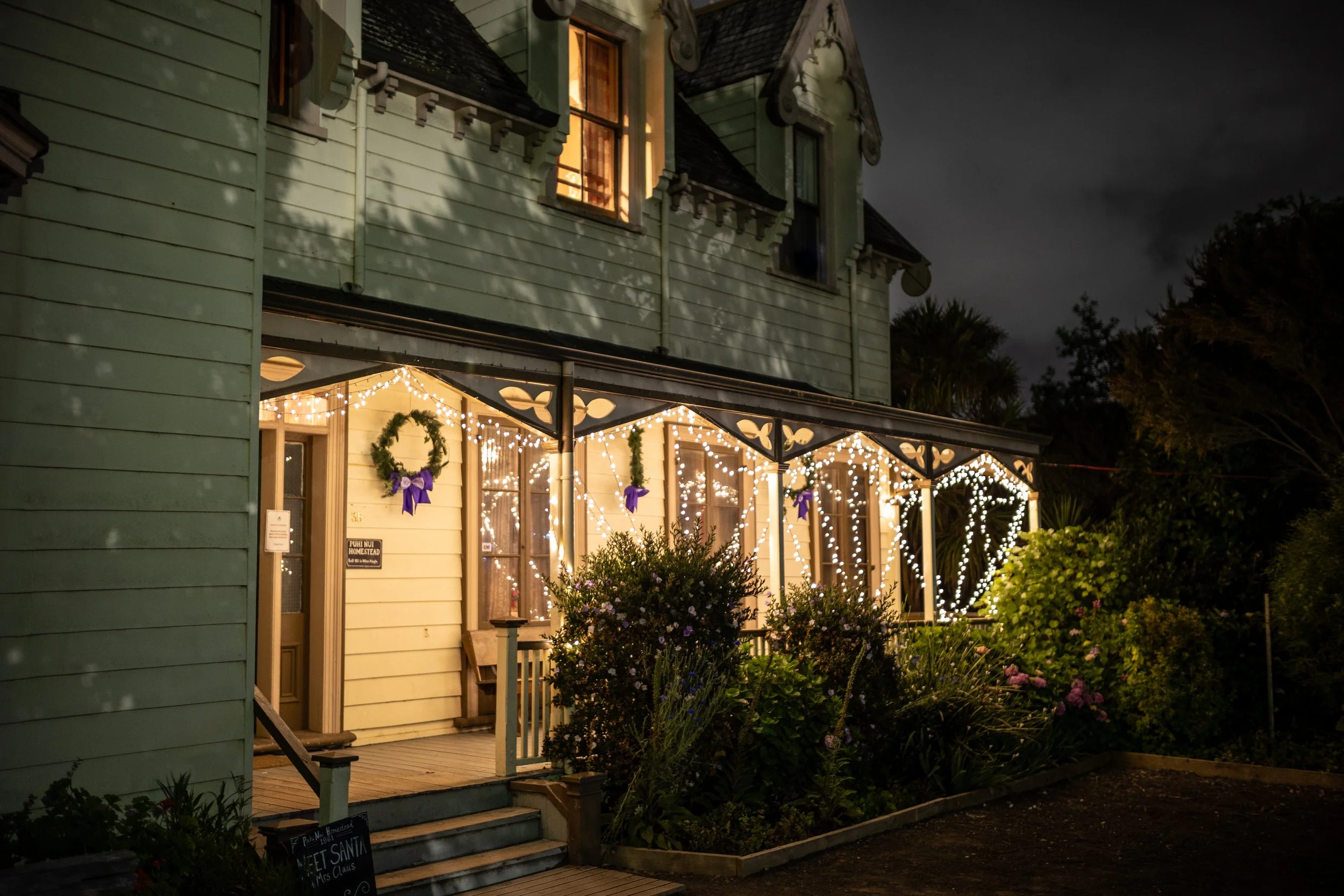 A brightly decorated house at night with Christmas lights and wreaths on the porch, surrounded by flowers and shrubs.