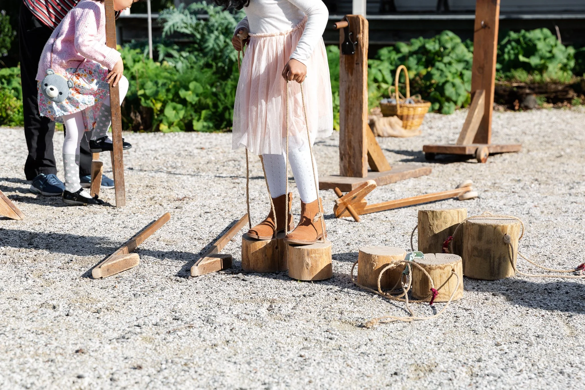 Children playing with wooden stilts and log stepping blocks on gravel outdoors.