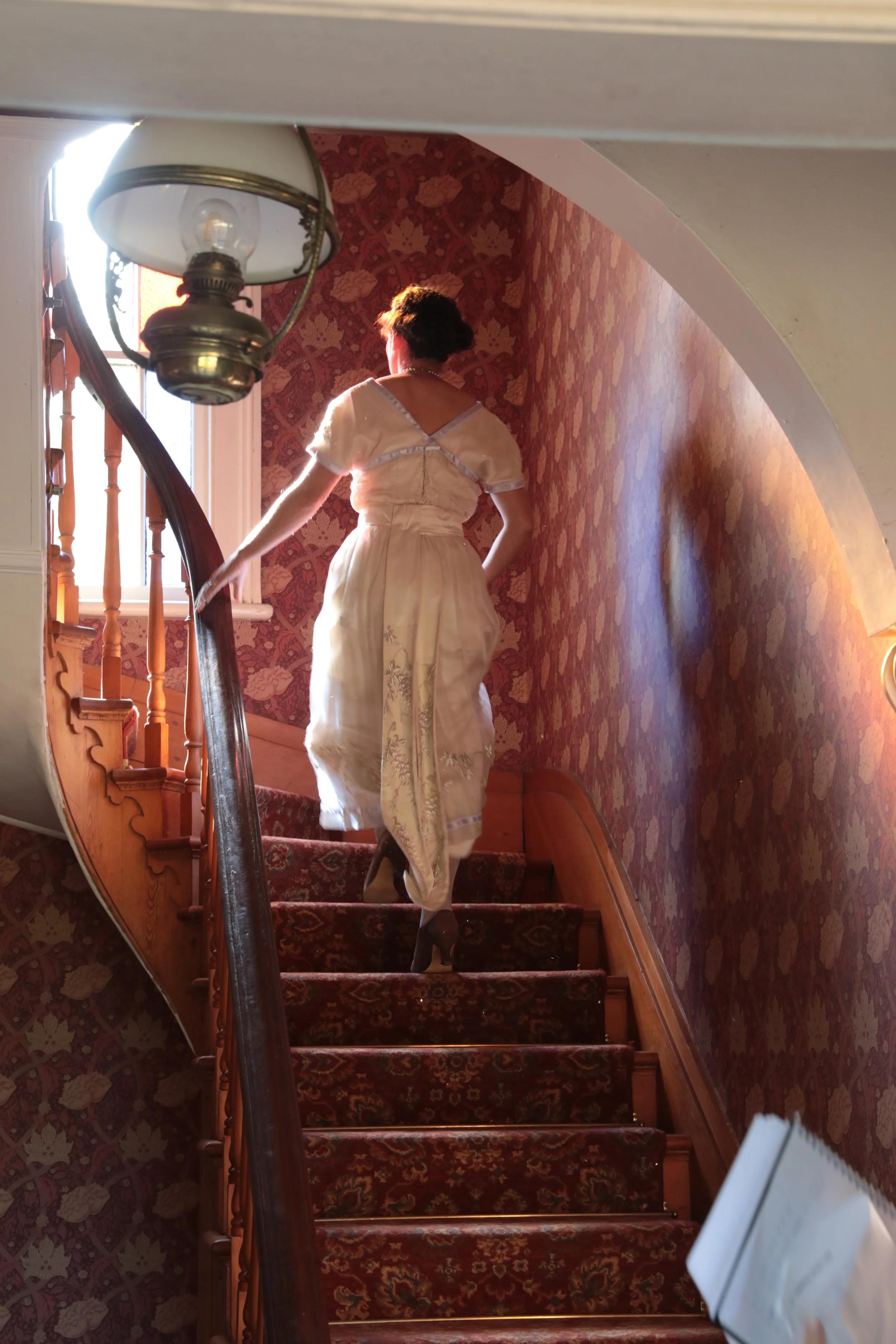Woman in a vintage cream-colored dress walking up a carpeted staircase with wooden railings, near a window with sunlight, and an hanging lamp.