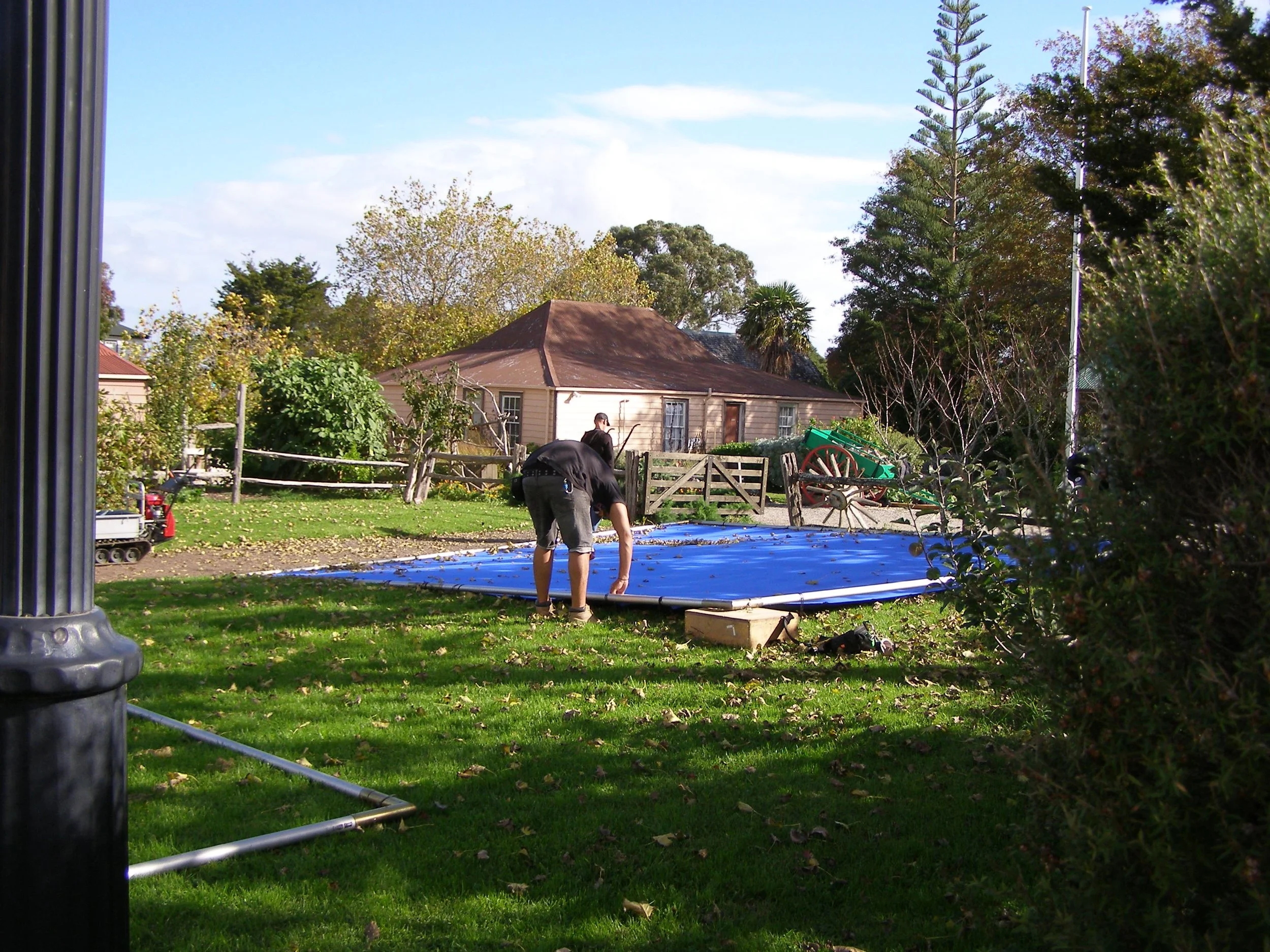 People setting up a blue above-ground swimming pool in a backyard yard with green grass, trees, and a house in the background.