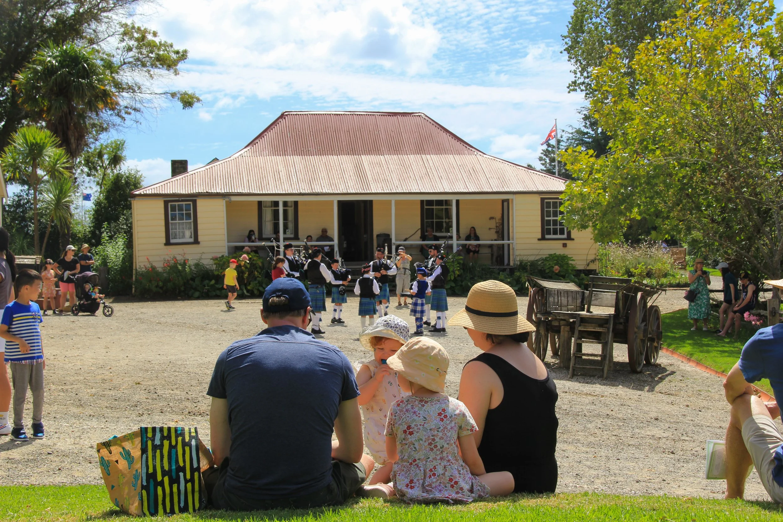 Group of people sitting on grass watching a bagpipe band perform in front of a yellow house with a red roof. Children and adults are gathered around; some standing, some sitting, some playing in the yard. Trees and blue sky are visible.