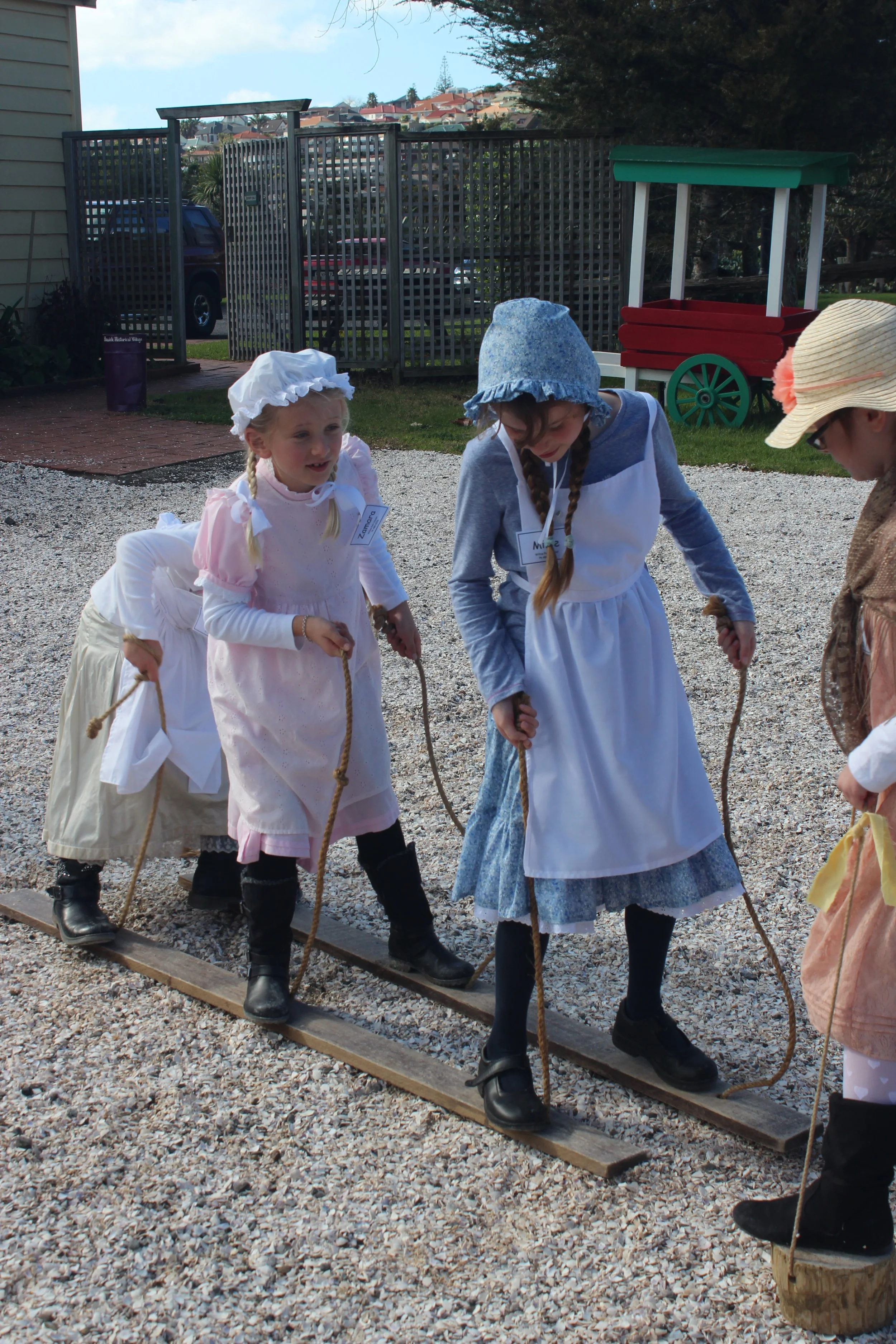 Children dressed in historical clothing walking on a wooden balance beam outdoors.