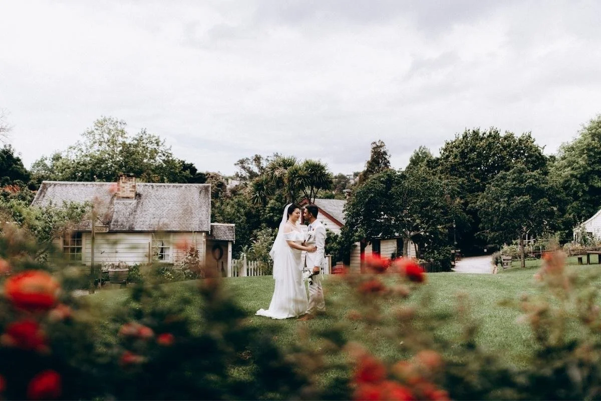 A bride and groom holding hands in a grassy garden with houses, trees, and cloudy sky in the background.