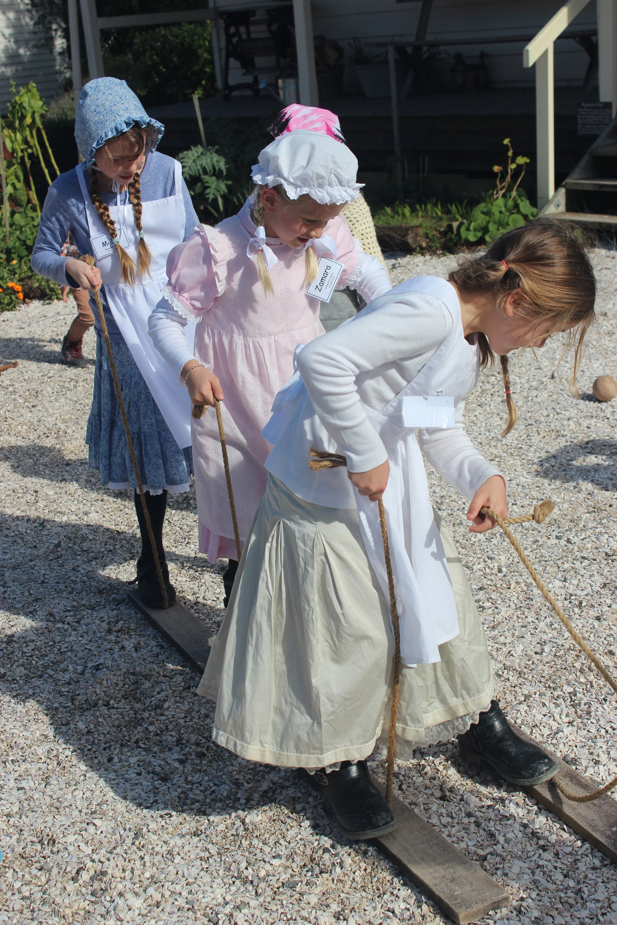 Four young girls dressed in historical clothing participate in an outdoor activity, standing on a wooden boardwalk and holding a rope, with a garden and steps in the background.