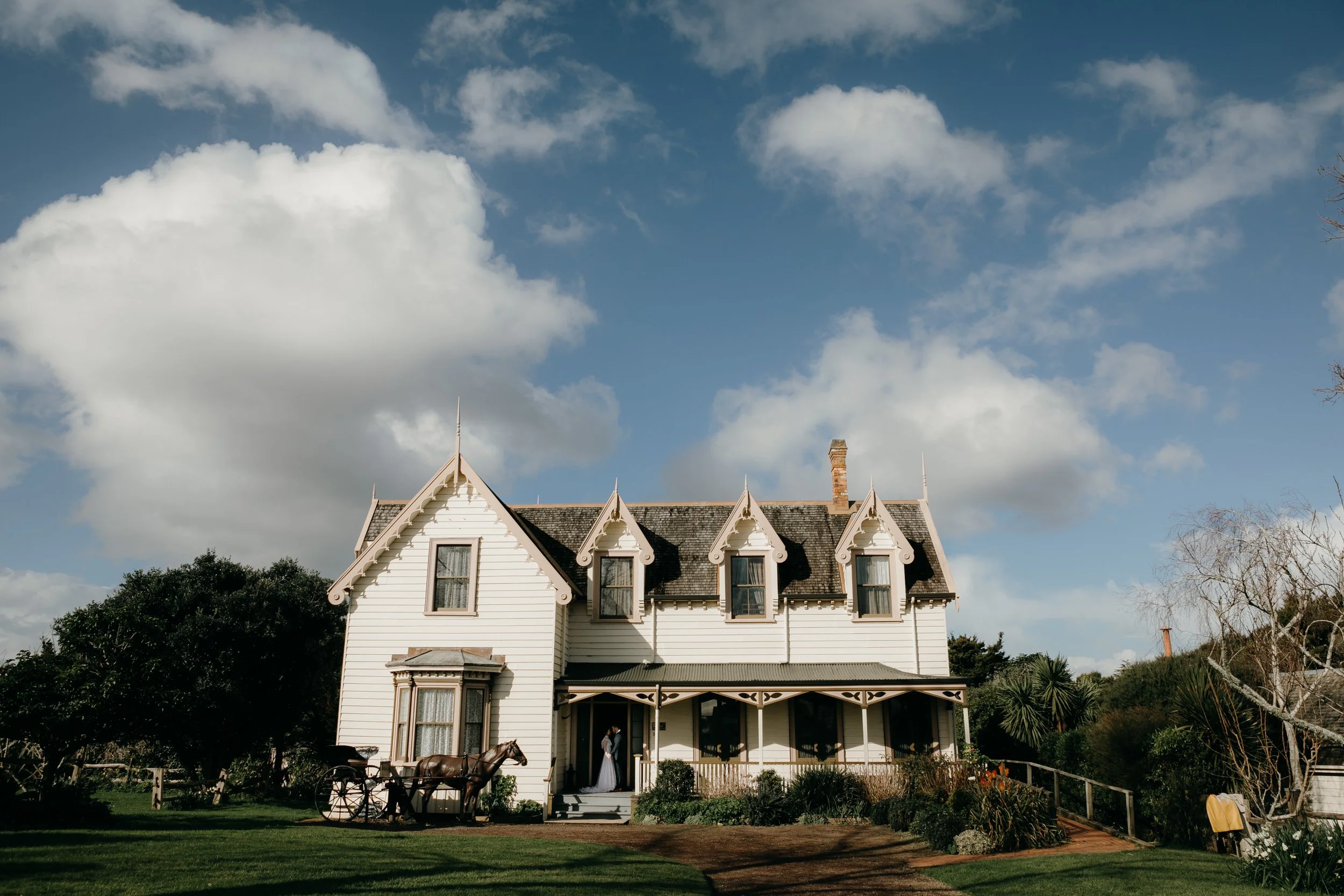 A white Victorian-style house with decorative trims, a carriage in front, surrounded by greenery, under a blue sky with clouds.