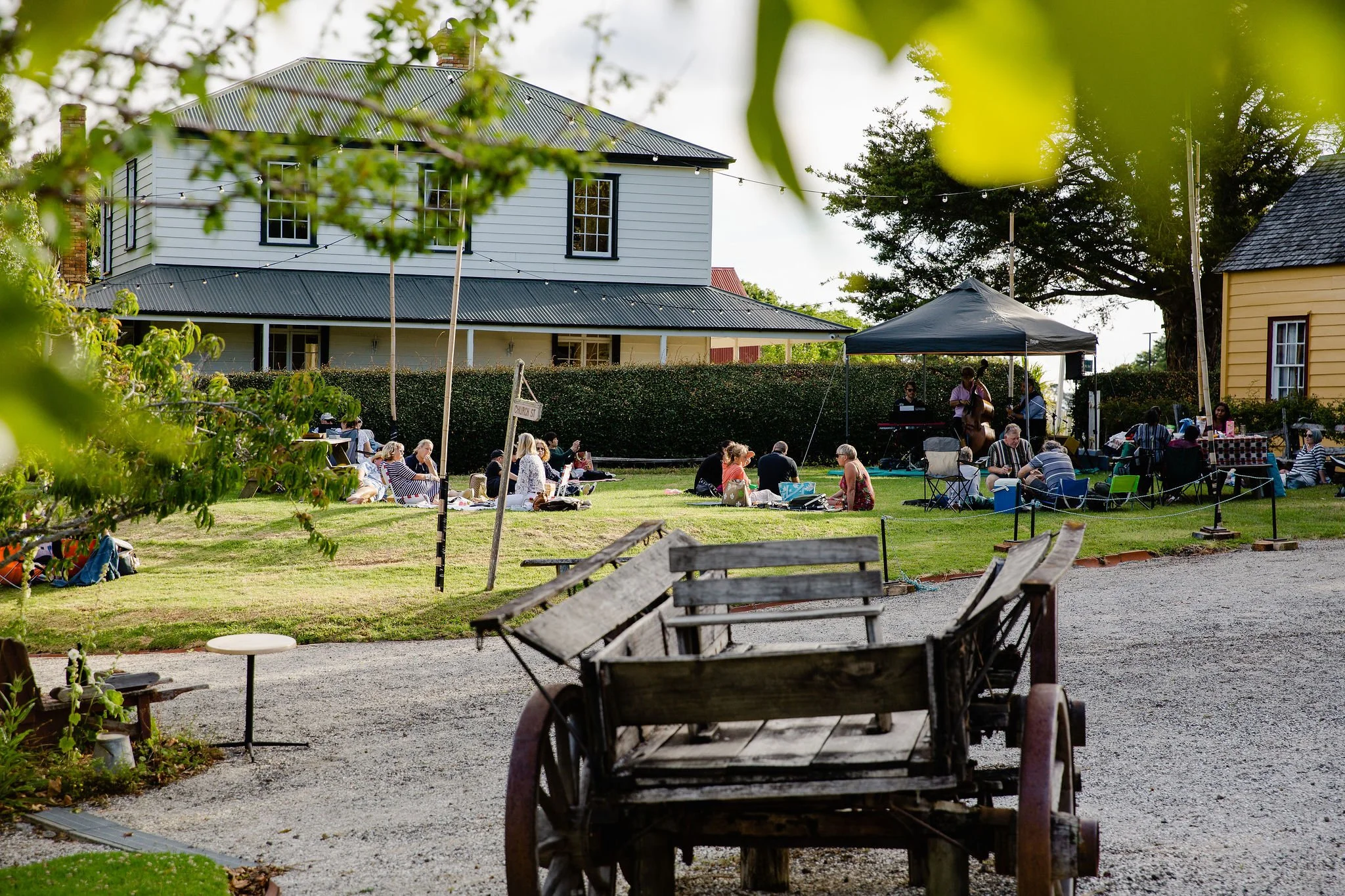 People enjoying a picnic and outdoor music performance on a lawn in front of a wooden house, with a canopy and an old wooden cart in the foreground.