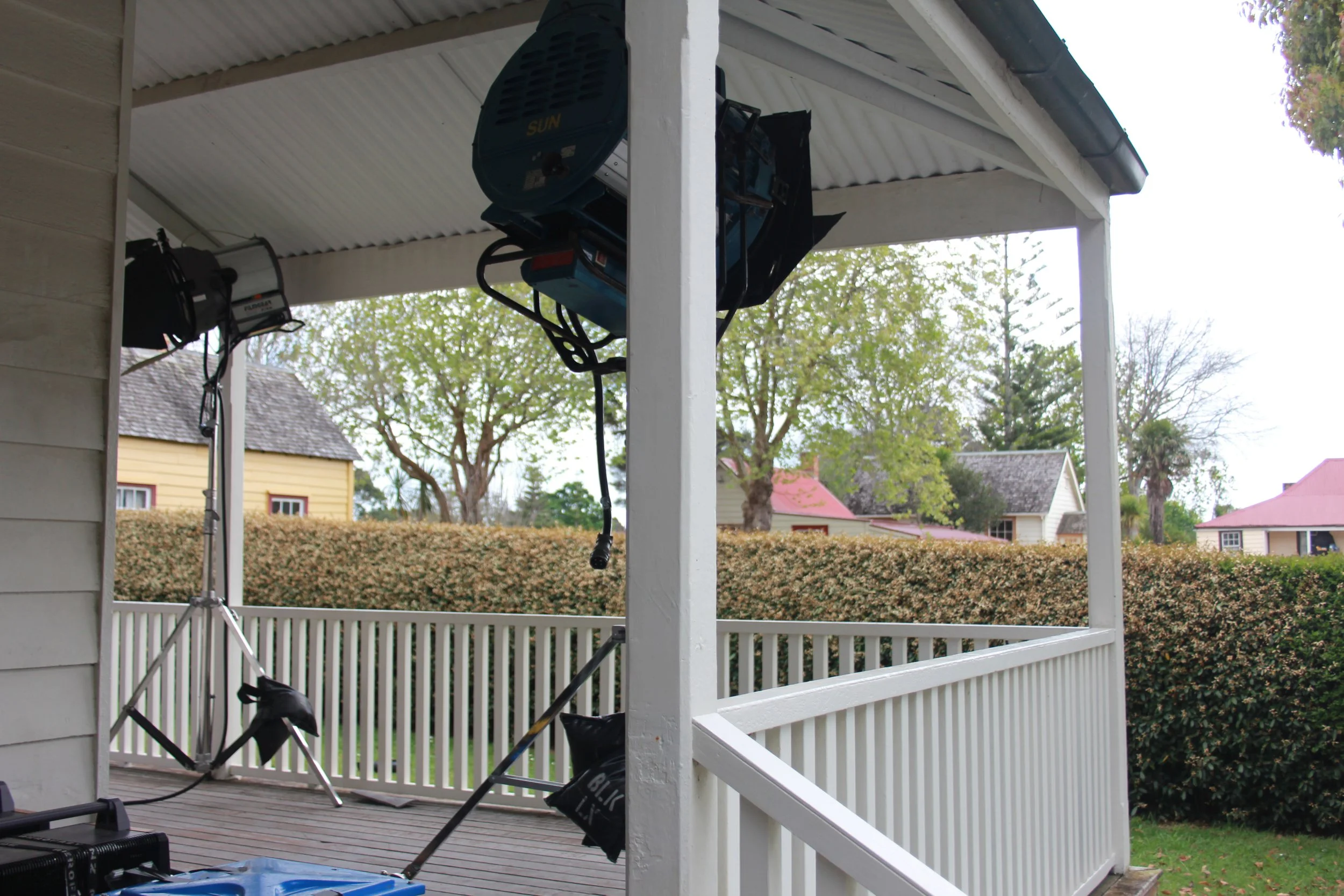 Back porch with studio lighting equipment, including a large light with a softbox, a tripod, and other gear, overlooking neighboring houses and trees.