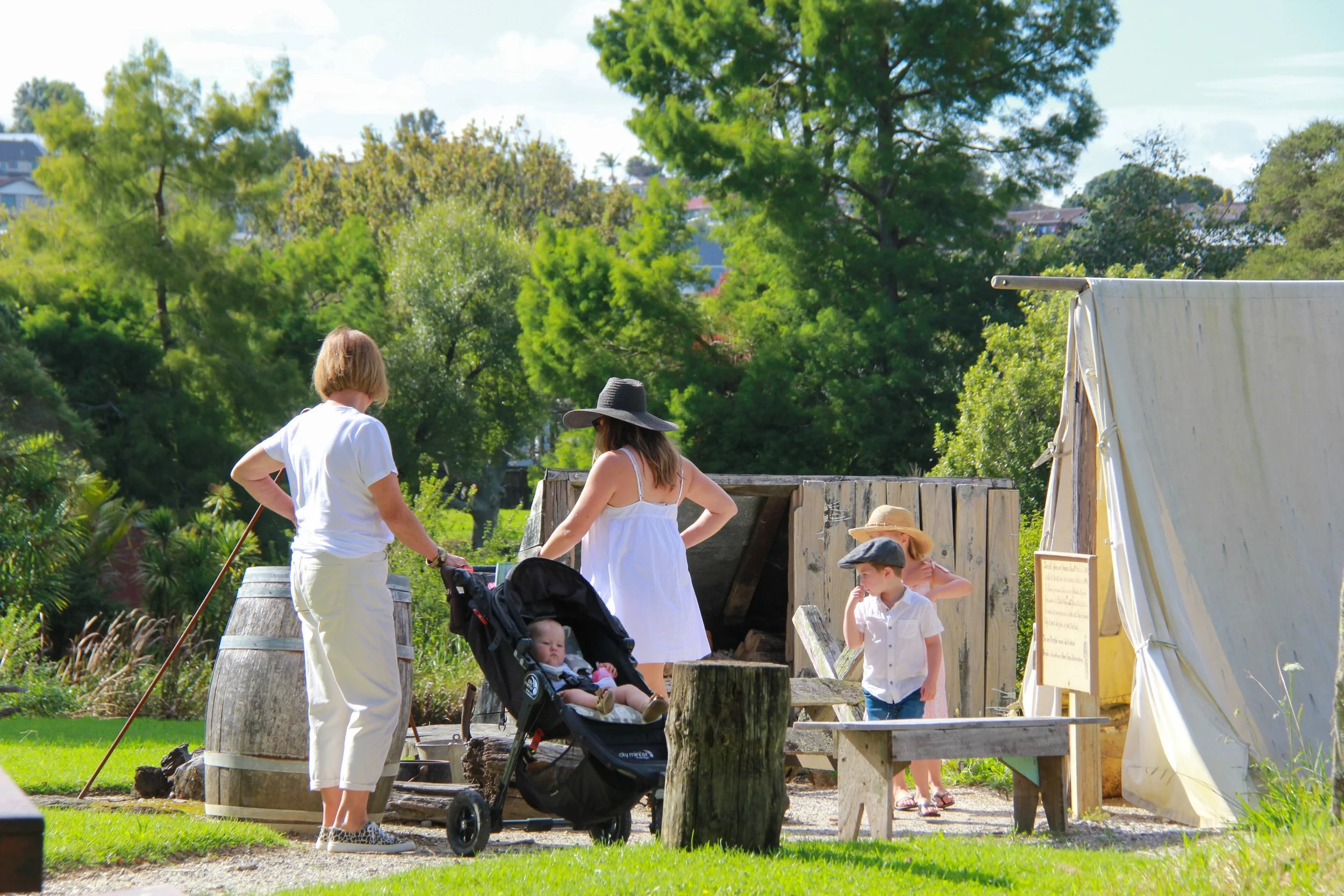 Family and children at an outdoor historical reenactment or themed event, with a tent, wooden barrels, and lush green trees in the background.