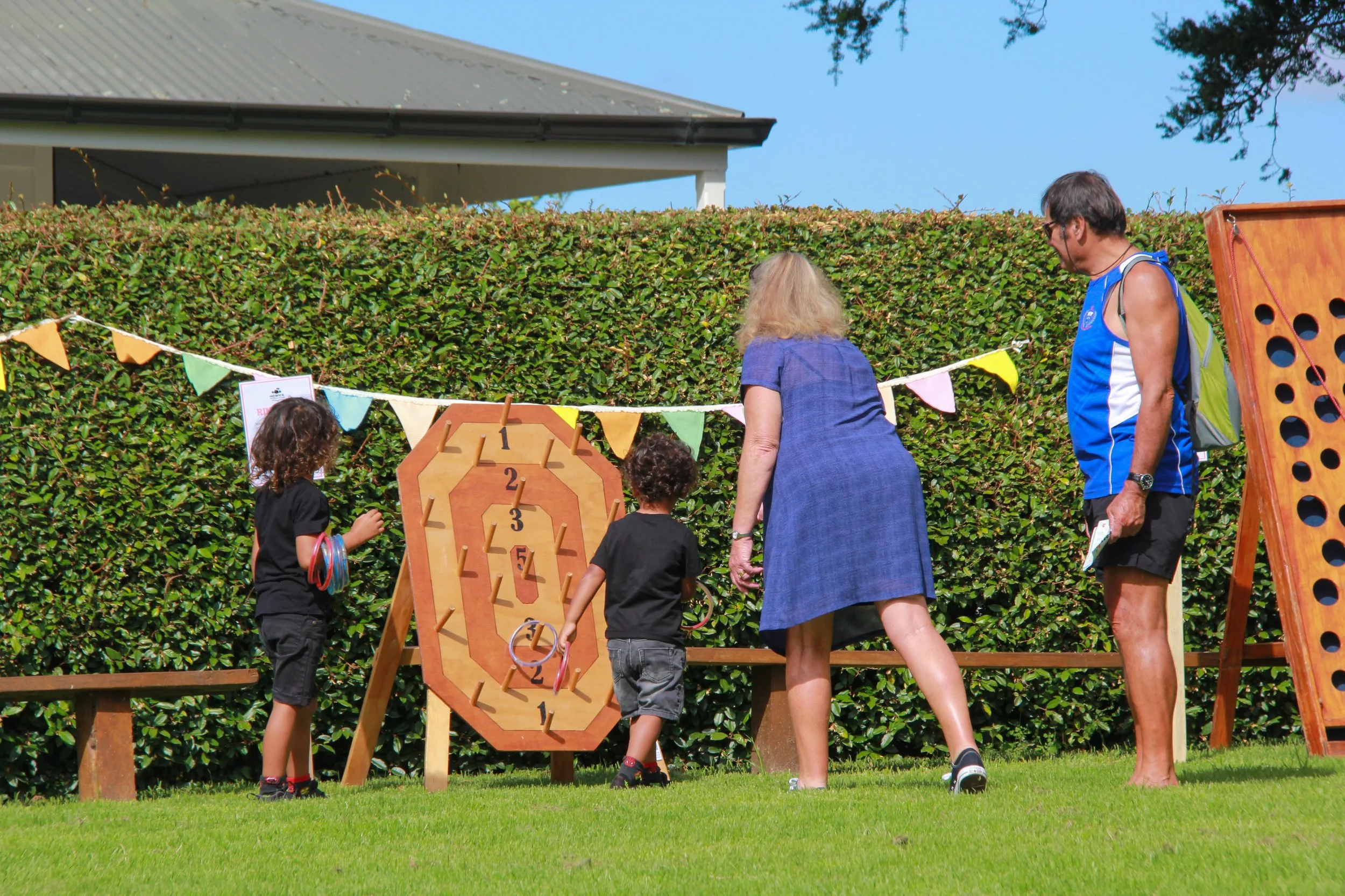 Children playing a ring toss game outdoors with two adults supervising, set against a green hedge and a house with a grey roof.