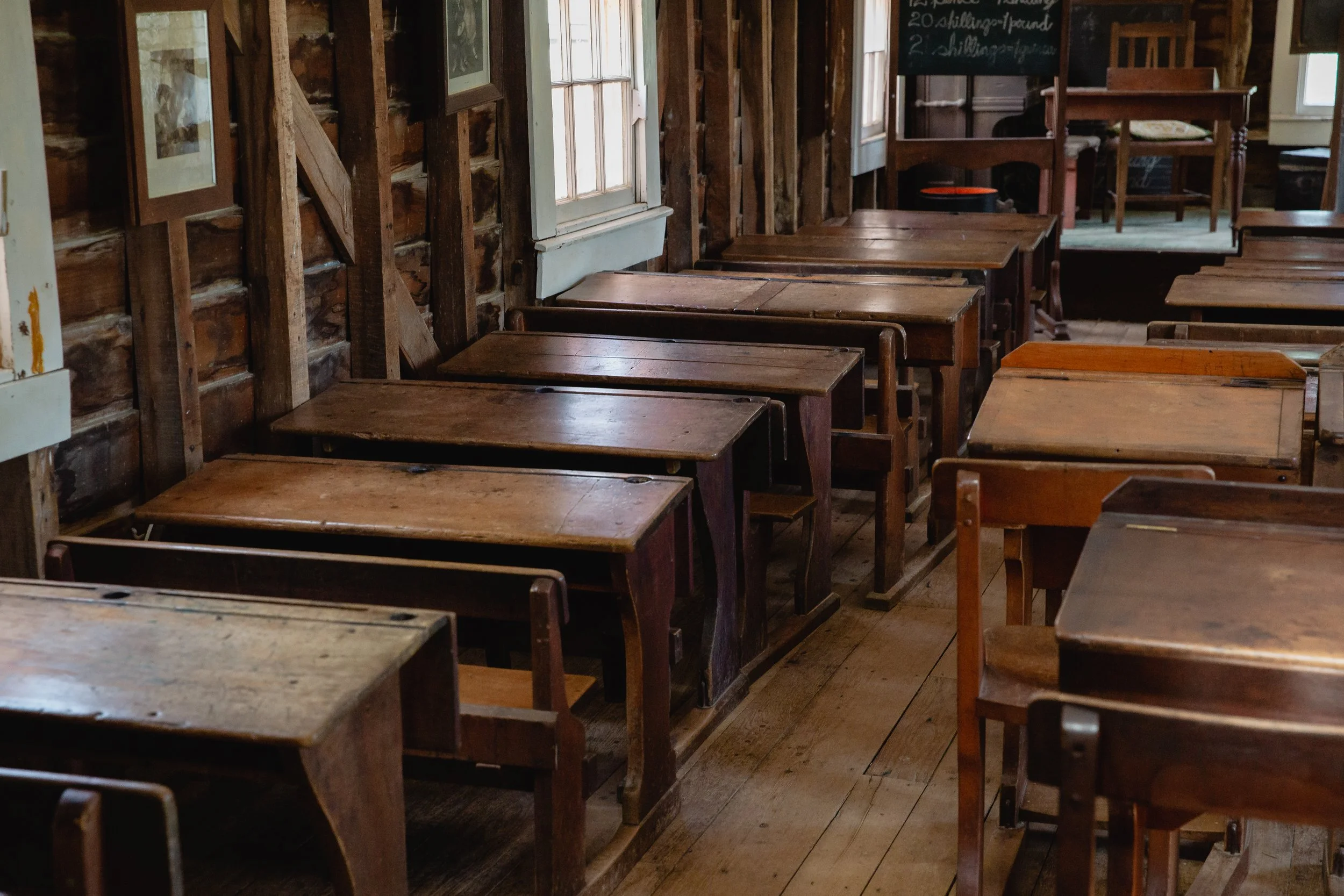 Interior of a rustic wooden classroom with vintage desks and chairs, wood-paneled walls, and a chalkboard in the background.