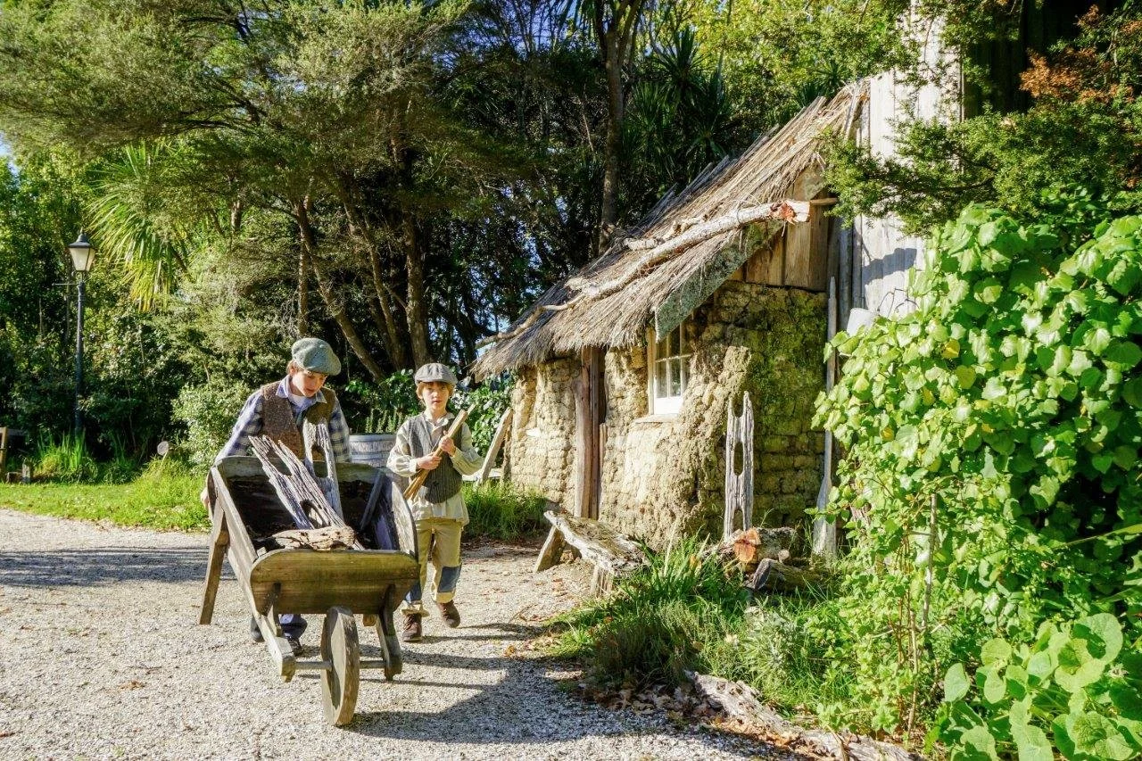 Two people in vintage clothing with a wooden wheelbarrow outside a rustic thatched cottage in a garden setting.