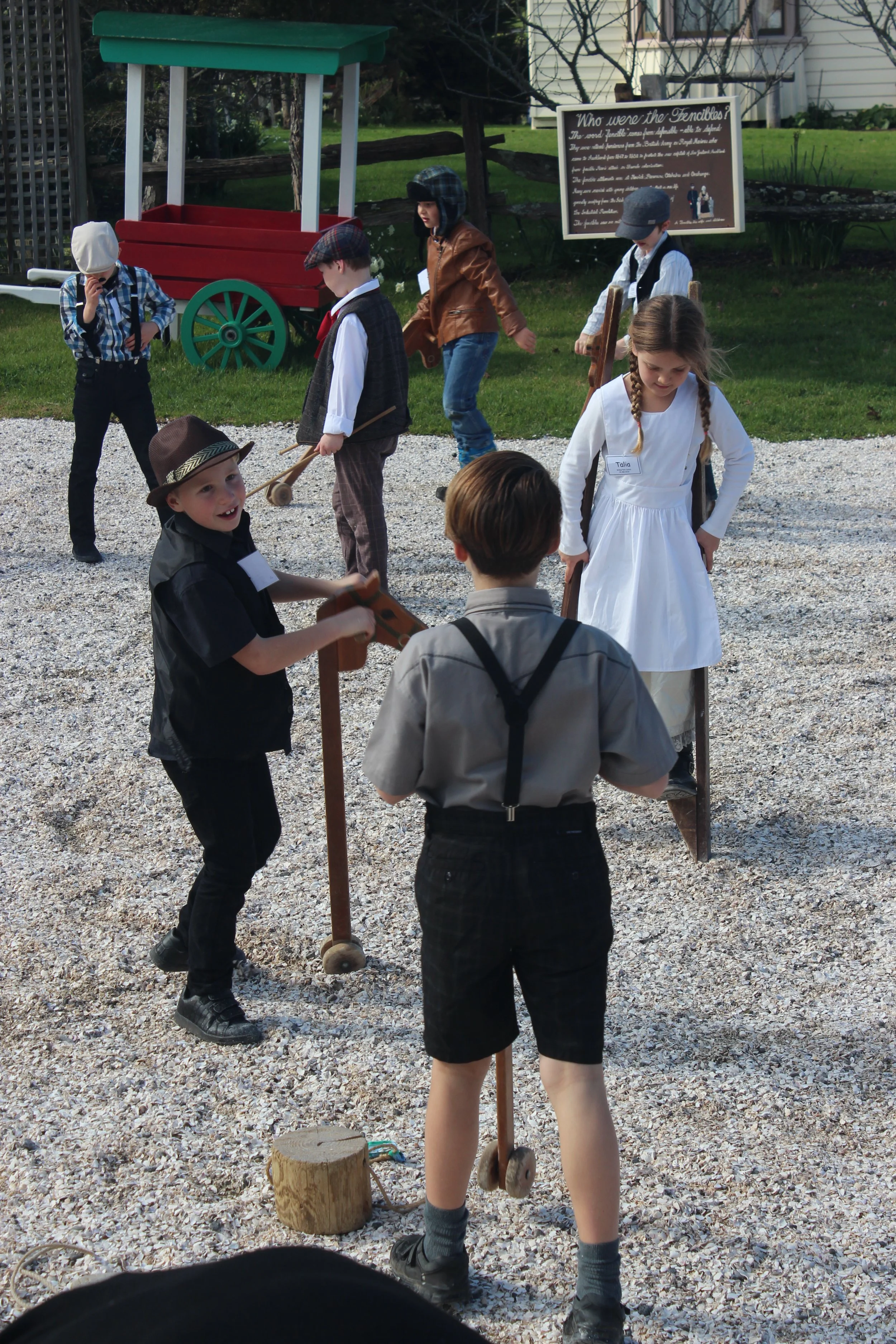Children playing a game on scooters outdoors, dressed in period costumes, with some adults observing. A sign about the Fencibles stands in the background.