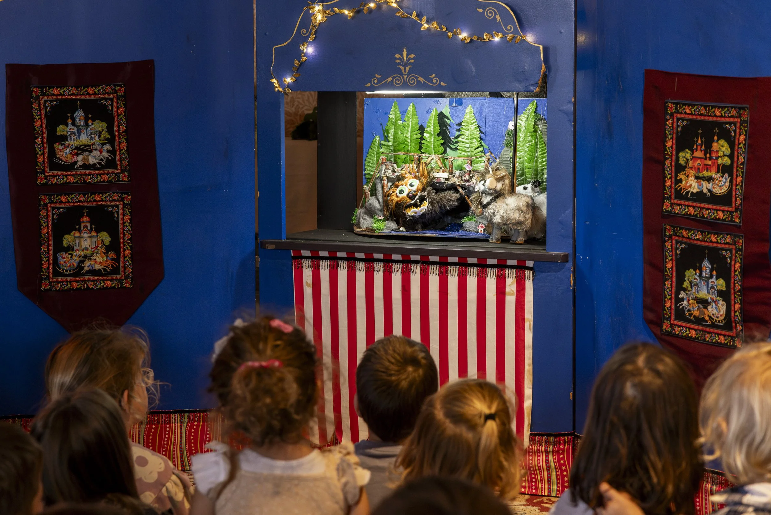 Children watching a puppet show with a puppet horse, lion, and trees on stage, decorated with string lights.