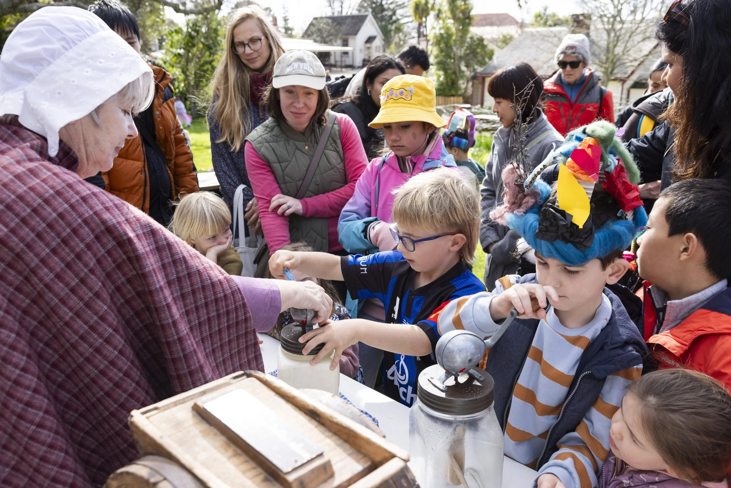 Group of children and adults at an outdoor educational event, observing a woman demonstrating something with jars on a table, surrounded by trees and houses.