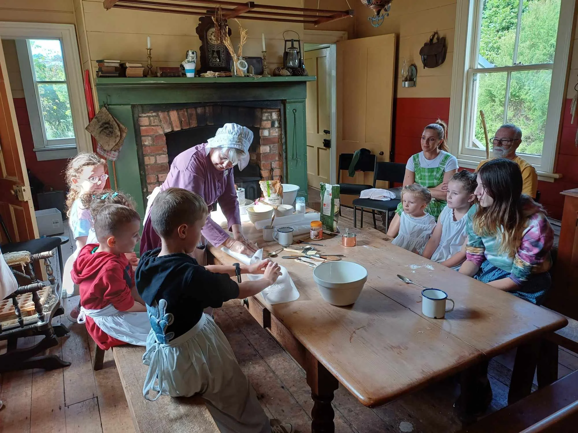 A group of children and adults gathered around a large wooden table in a rustic kitchen with wood-paneled walls and windows, watching a woman dressed in a purple apron and chef hat prepare food.