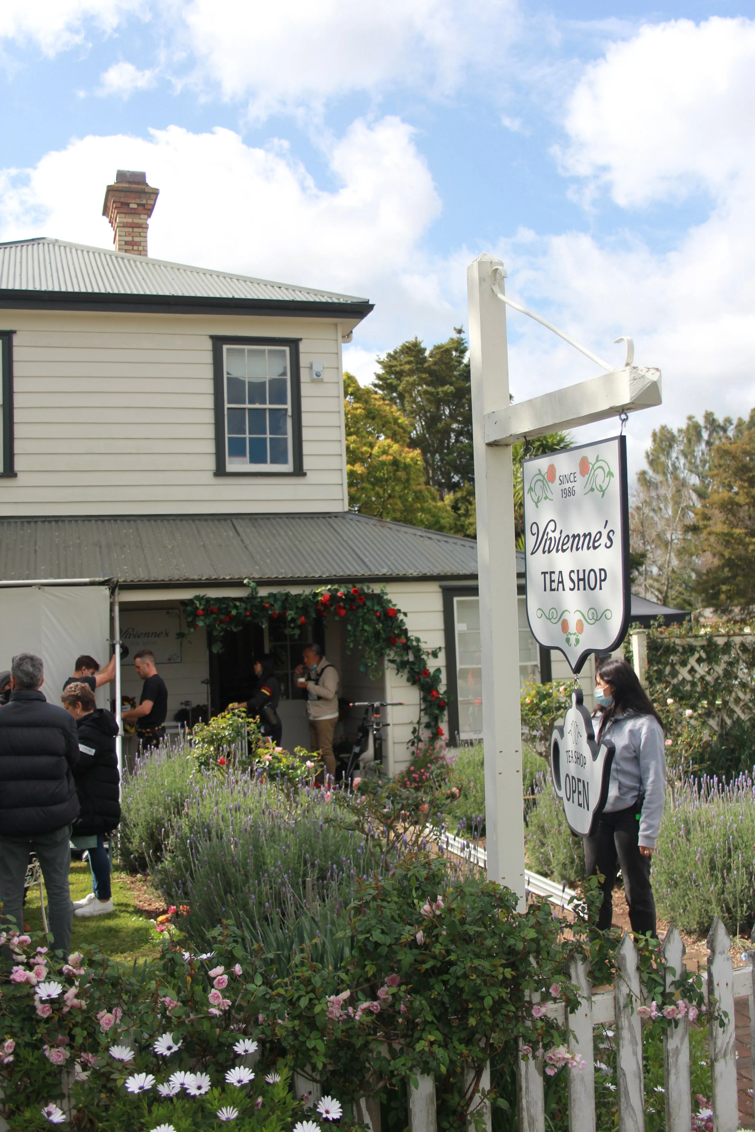 People standing outside Vivienne's Tea Shop, which is decorated with a floral arch and surrounded by pink and purple flowers on a sunny day.