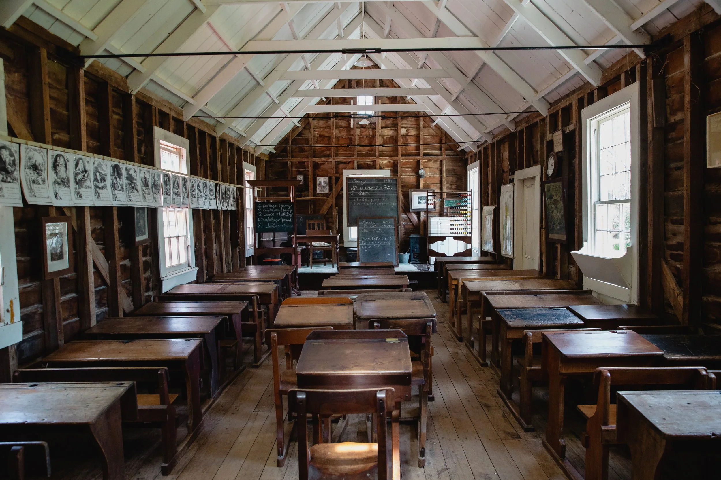 An old-fashioned classroom with wooden desks arranged in rows, large windows allowing natural light, and a chalkboard at the front with handwritten notes.