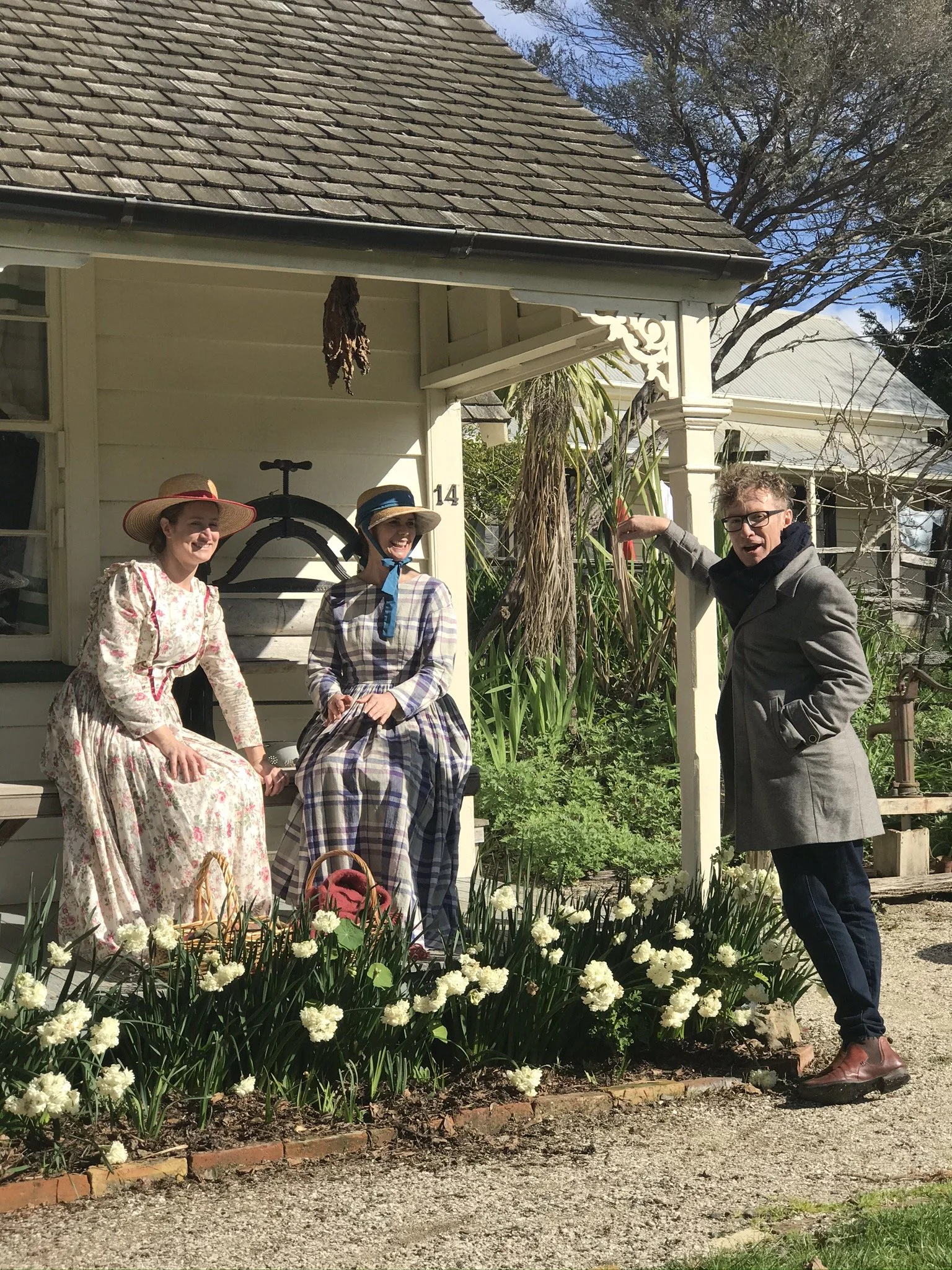 Two women dressed in vintage clothing and a man in modern clothing in front of a house with garden flowers.
