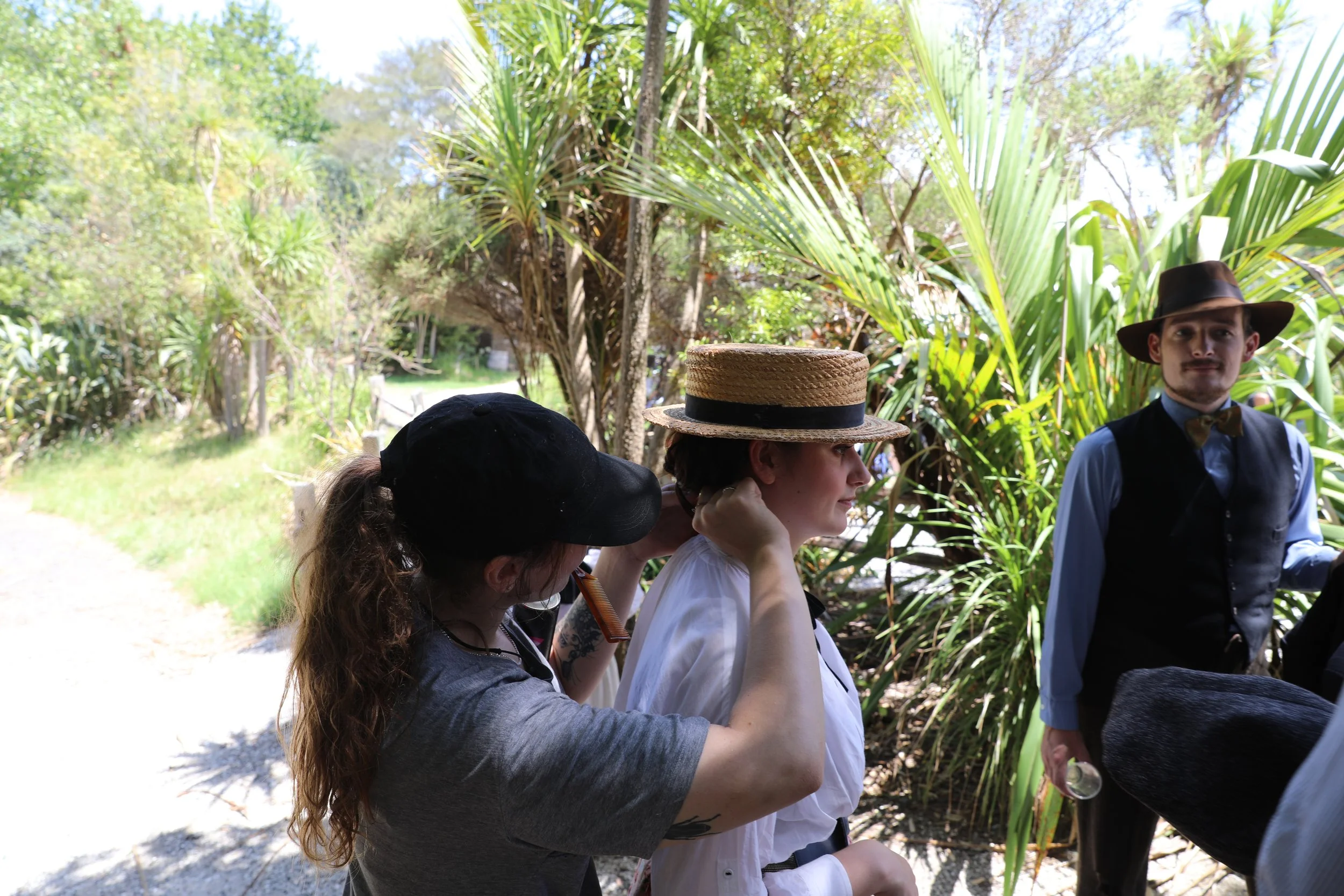 Three people, two women and one man, wearing vintage or retro clothing, are standing outdoors among lush green tropical plants. The woman in the middle is adjusting her straw hat, and the man on the right is wearing a wide-brimmed hat and vest, holdi