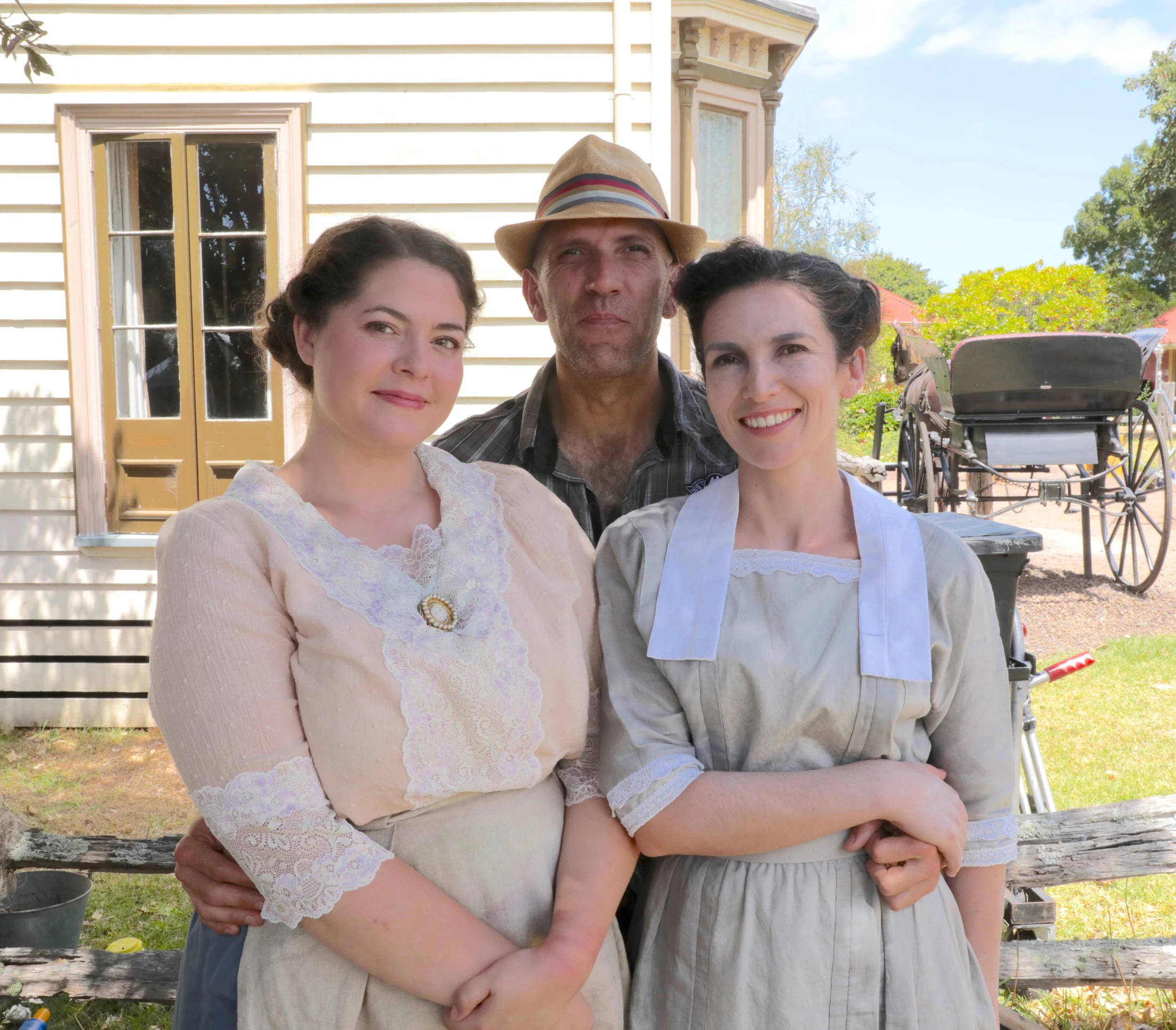 Three people dressed in 19th-century clothing standing outdoors in front of a house and a wagon.