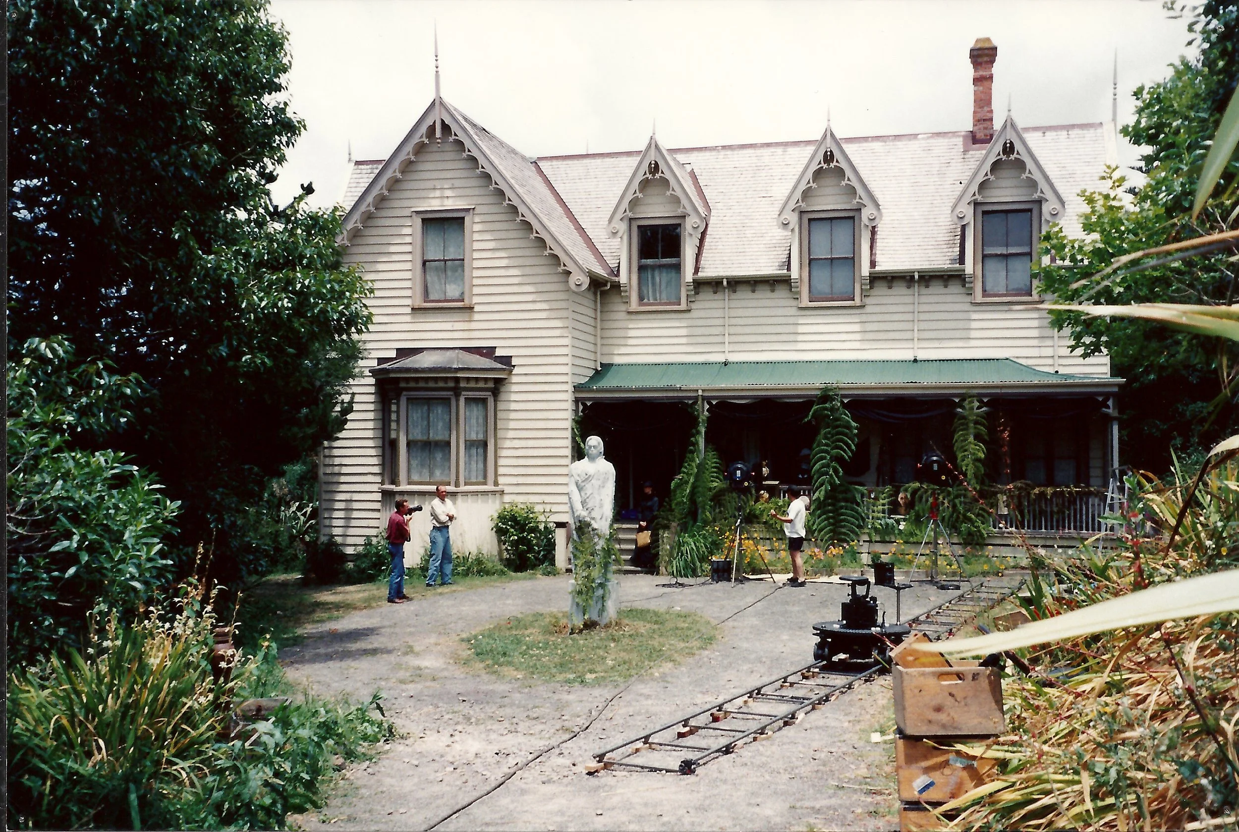 Film set scene in front of a large Victorian-style house with a green roof. Crew members, film equipment including a camera on a track, and a small statue planted in front of the house are visible.