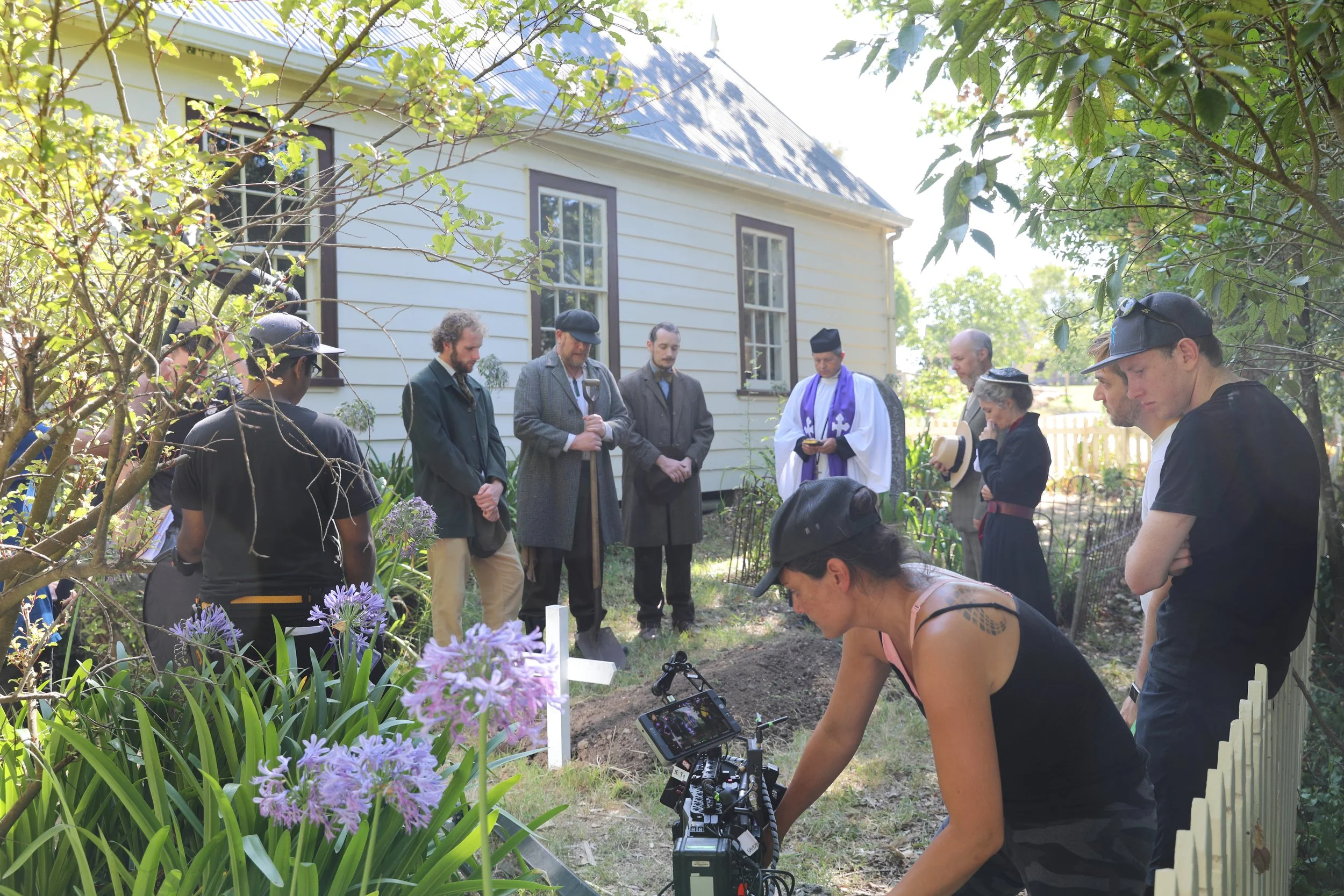 Group of people participating in a memorial or ceremony outdoors near a white house, with a woman filming with a camera, surrounded by trees and flowering plants.