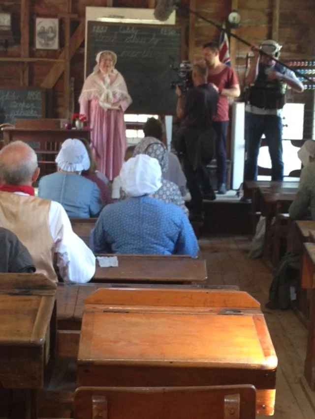Reenactment of a historical scene with actors in period costumes performing for an audience in a rustic setting, with vintage decor and wooden furniture.