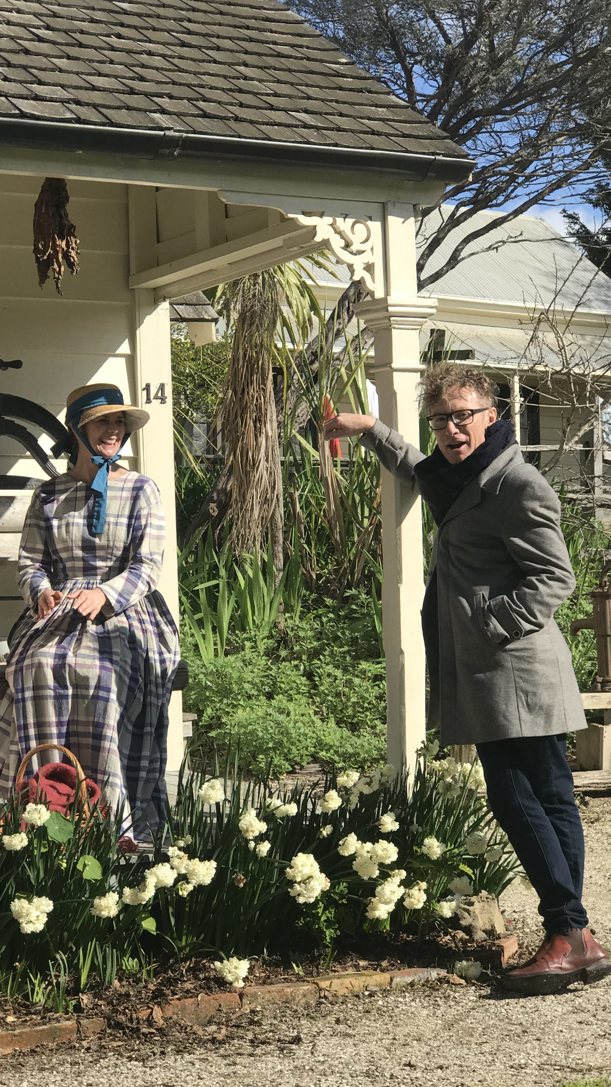 A woman dressed in vintage clothing and a wide-brimmed straw hat with a blue ribbon, sitting on a small bench in a garden, smiling at a man in modern clothing and glasses, pointing at plants near a white wooden porch. White flowers are in the garden,
