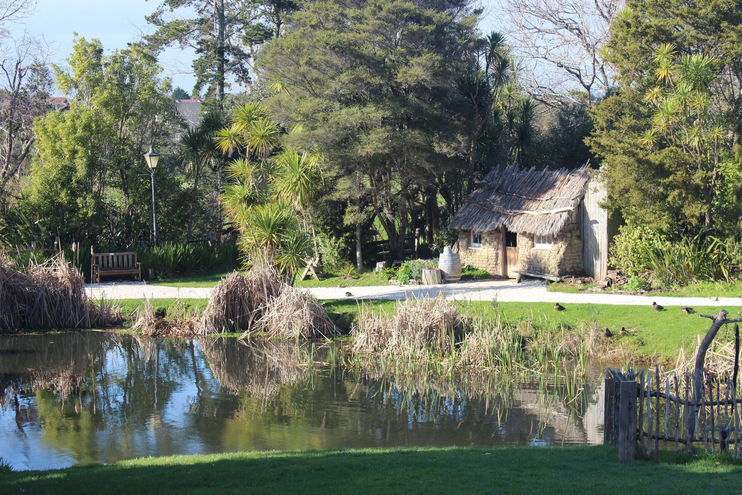 A small rustic hut with a thatched roof beside a pond surrounded by trees and greenery, with a wooden bench, lamppost, and ducks on the grass.