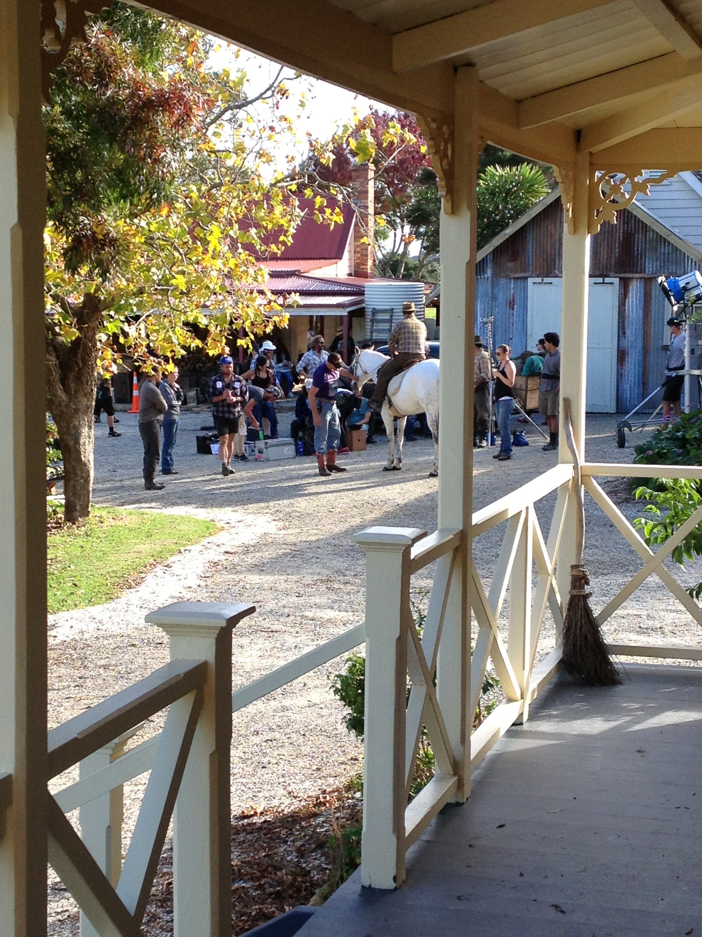 People gathered outside a rustic building with a horse and a person riding it. The scene is viewed from a porch with a white railing, and there are trees and buildings in the background.