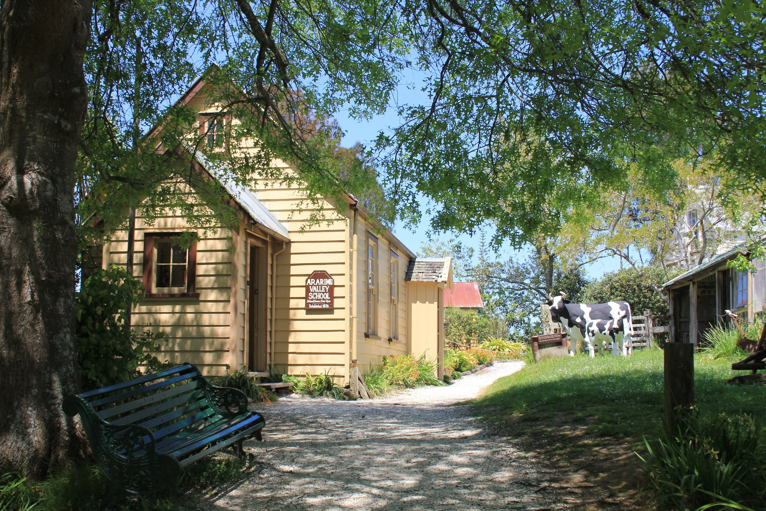 A sunny outdoor scene with a large tree and green leaves casting shade over a park area. There are two green benches, a small yellow building with a sign reading 'Ararimu Valley School,' and a farm scene with a black and white cow statue and various 