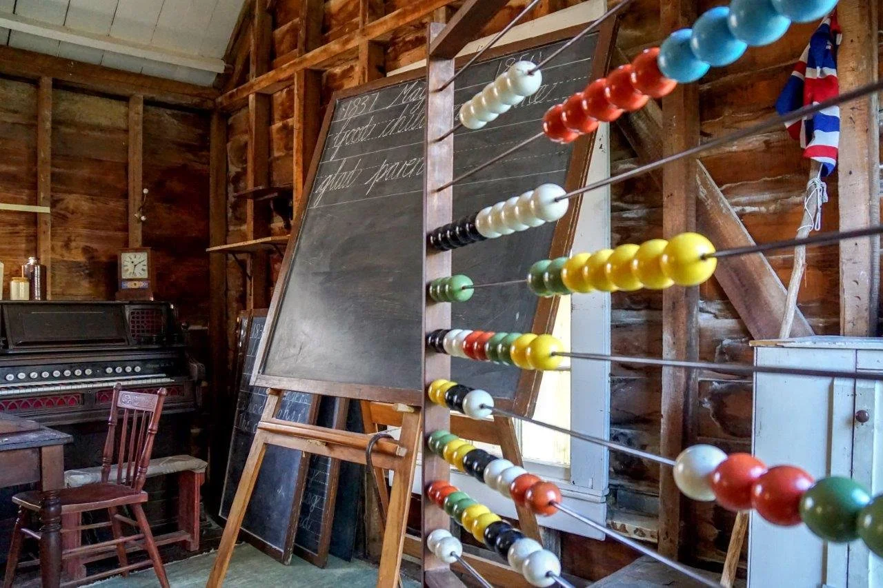 A vintage room with a blackboard, a wooden chair, an old piano, and a large set of colorful magnetic balls on thin wires, arranged on a stand. The room has wooden walls, some framed boards, and a clock. There is an American flag hanging near the wall